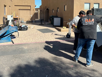 Two workers in a sunlit alley beside tan buildings, handling equipment near trash bins and a blue tarp.