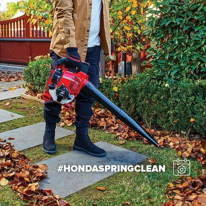A Man Is Blowing Leaves In A Garden With A Honda Leaf Blower — Ken Matthews Auto & Mower Centre In Ulladulla, NSW