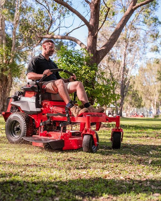 A Man Is Sitting On A Red Lawn Mower In A Park — Ken Matthews Auto & Mower Centre In Ulladulla, NSW