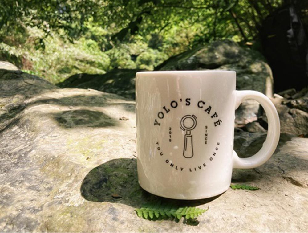 A white mug with the words yolo 's cafe on it is sitting on a rock.