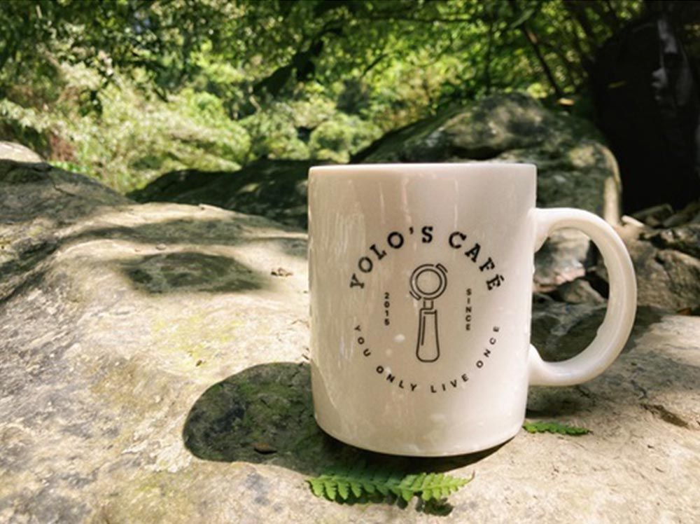 A white mug with the words yolo 's cafe on it is sitting on a rock.