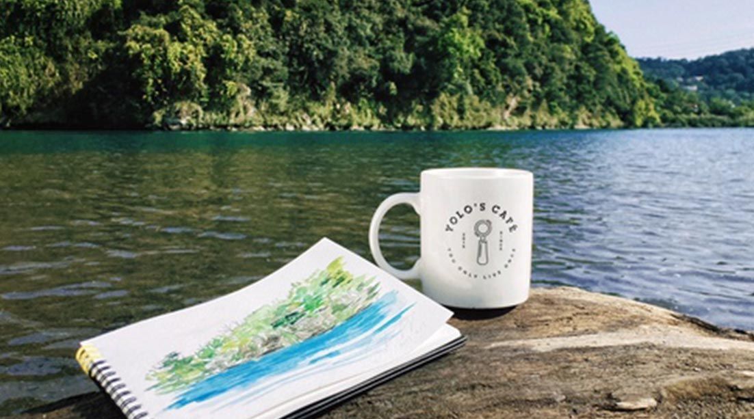 A mug and a notebook are sitting on a rock near a lake.
