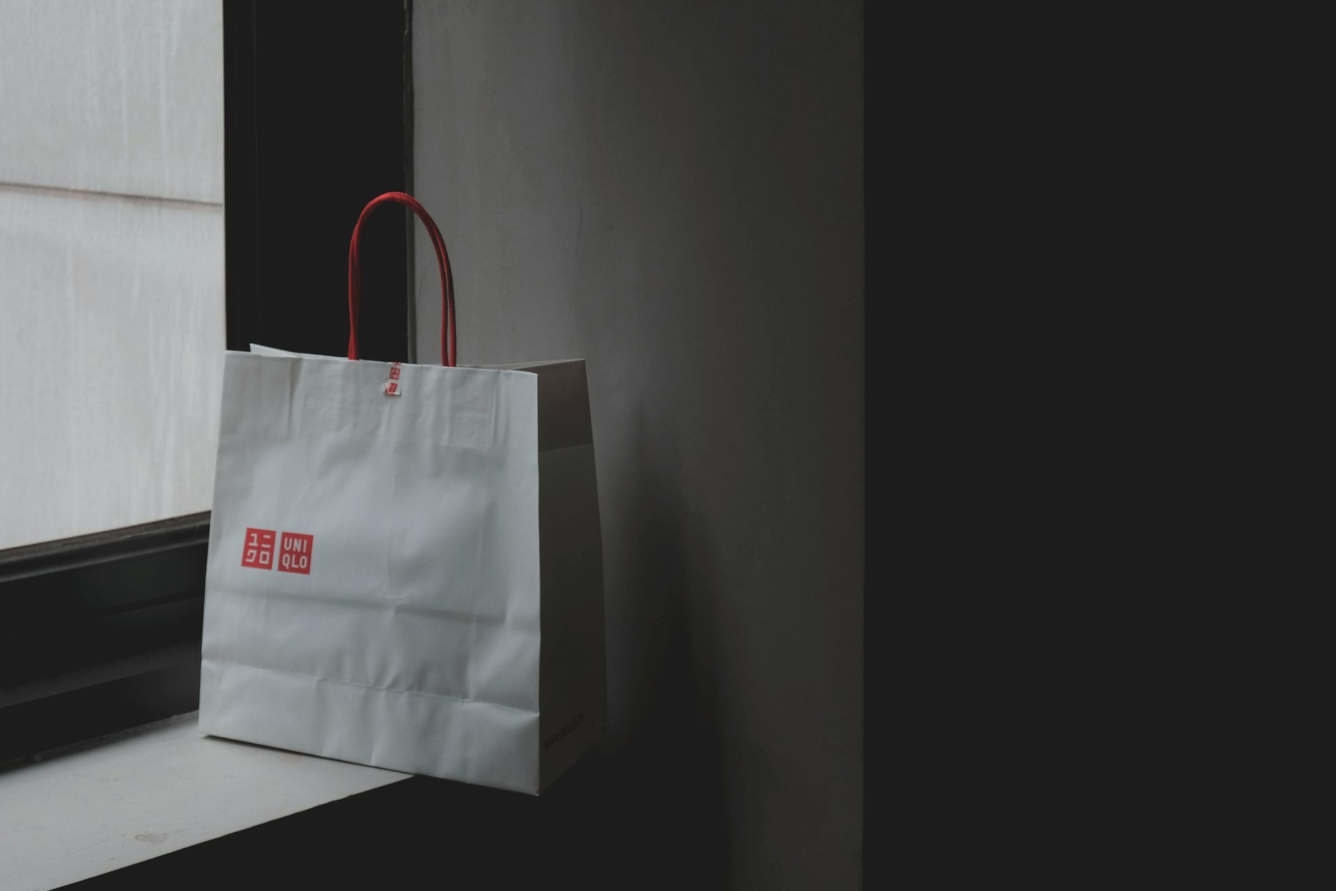 A white paper bag with a red handle is sitting on a window sill.
