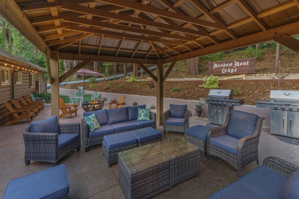 Patio area under a wooden pavilion featuring outdoor wicker furniture with blue cushions and a nearby grill station.