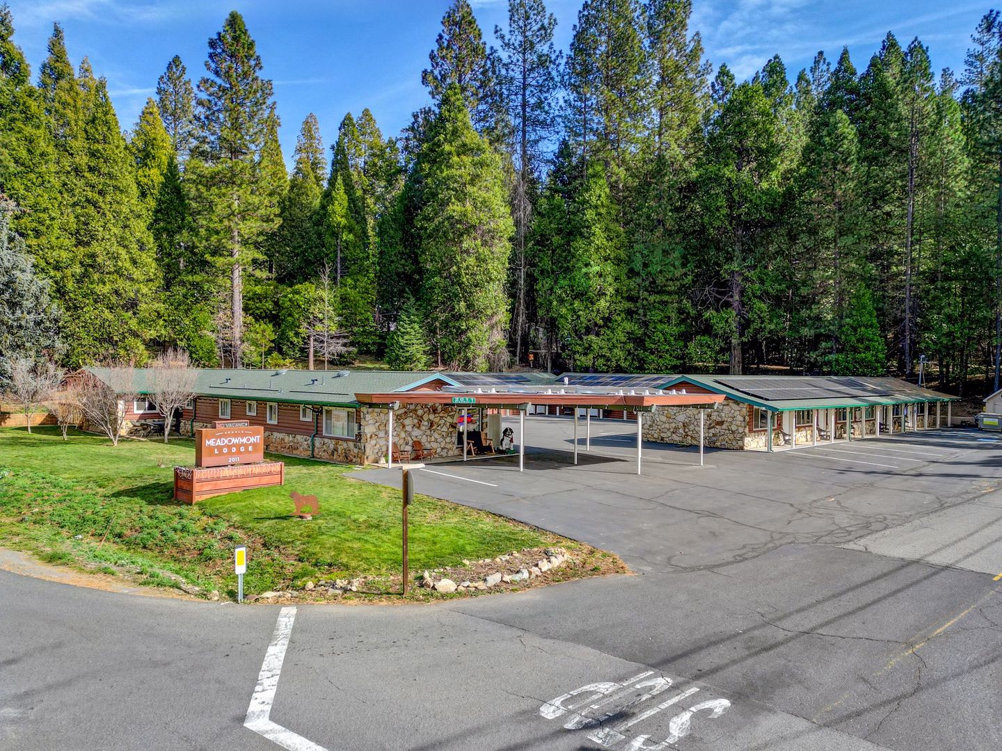 A single-story, stone-fronted motel surrounded by a dense pine forest, with a parking lot in the foreground.