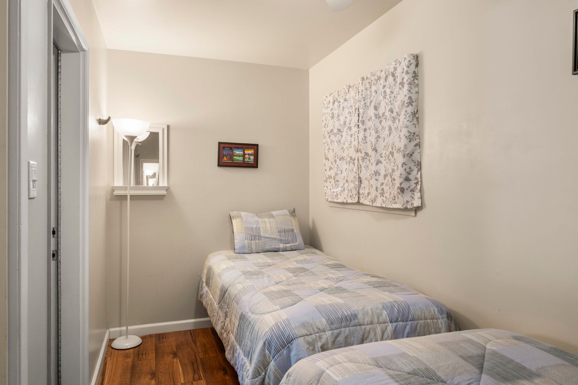 A small bedroom with two twin beds, hardwood floors, a floor lamp, and a patterned quilt set.
