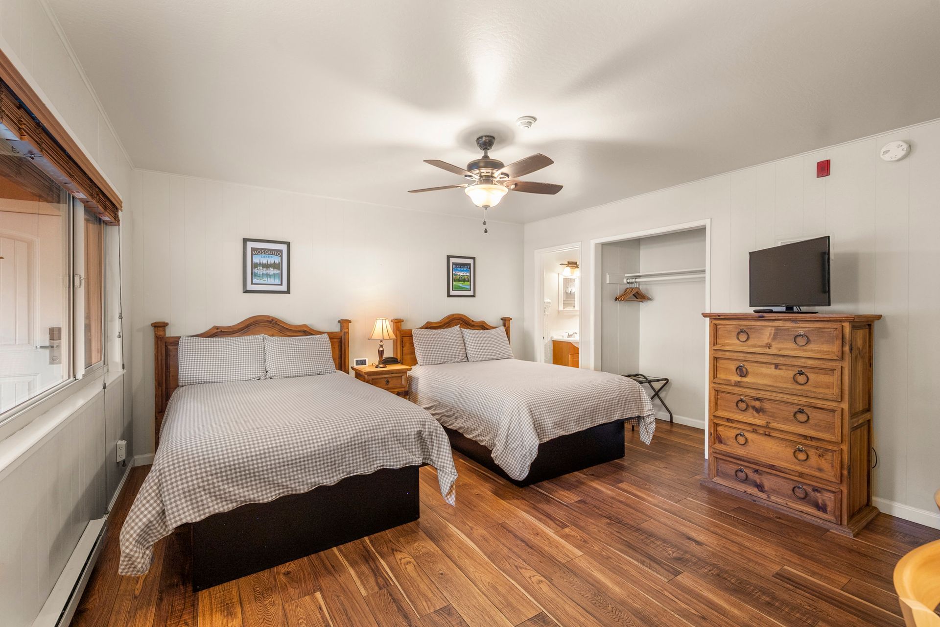 A bedroom with two beds, a wooden dresser holding a TV, a ceiling fan, and hardwood floors.