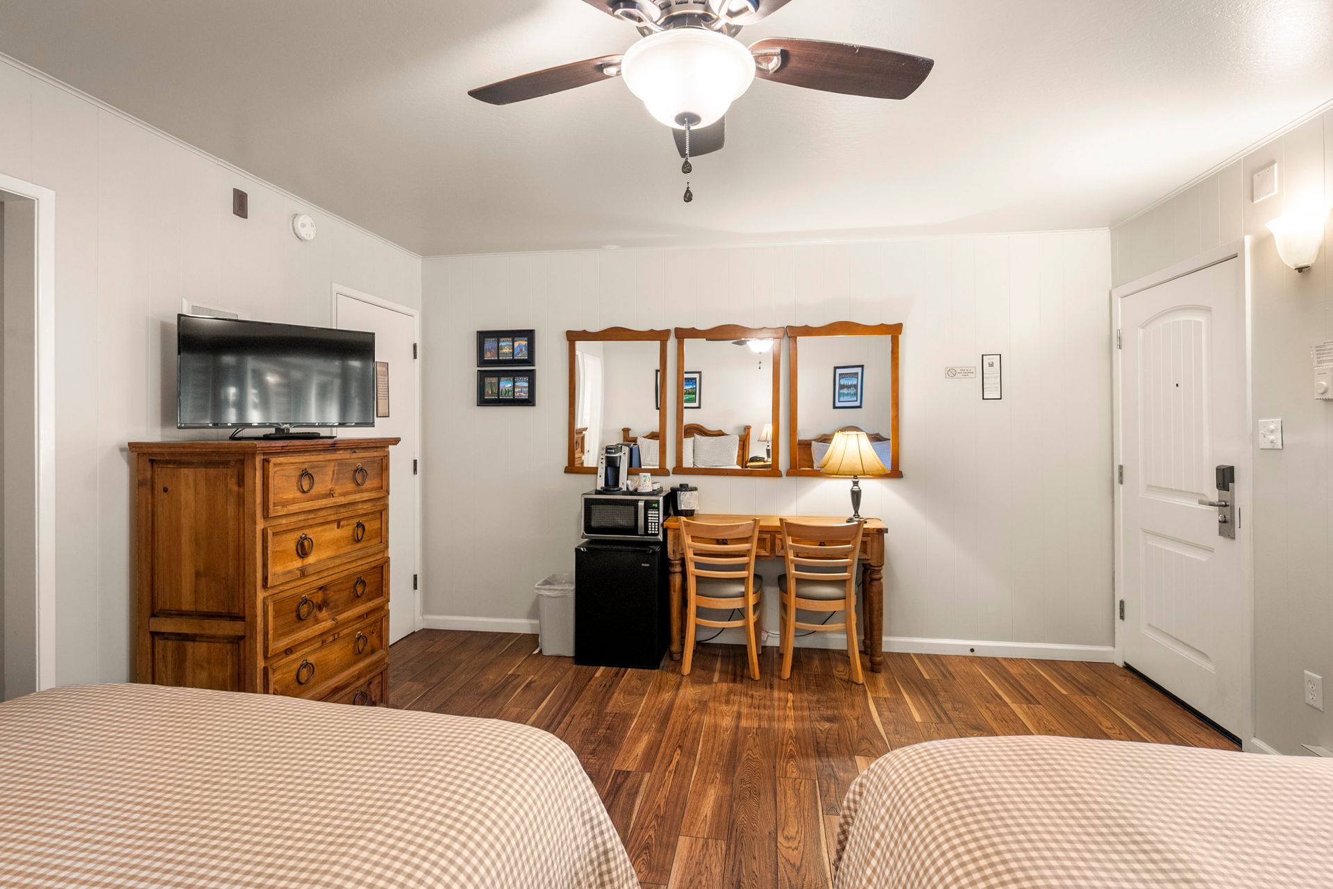 A hotel room with two beds, a wooden dresser, a television, a desk with two chairs, and a ceiling fan.