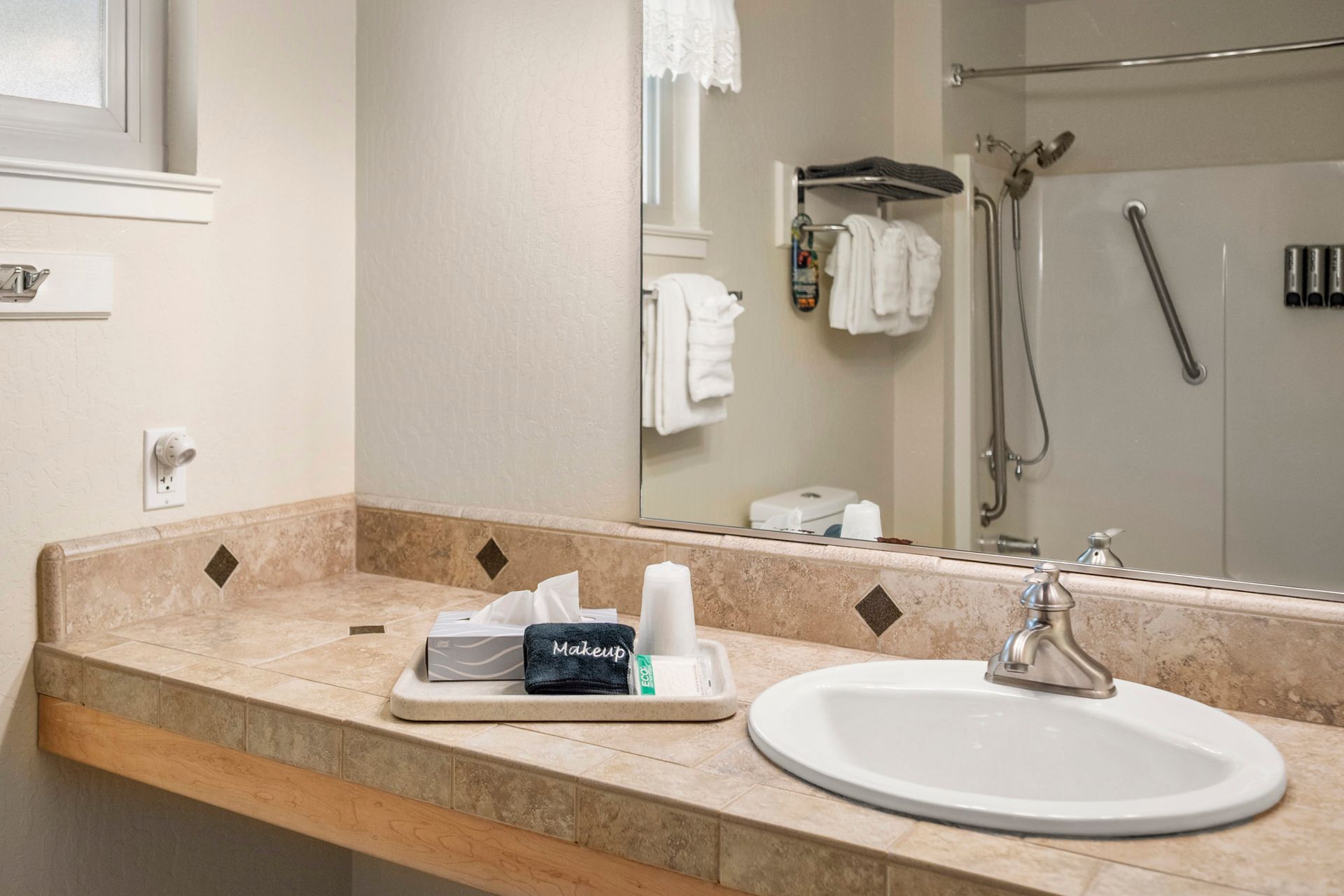 A hotel bathroom with a stone-topped vanity, white sink, mirror, and a walk-in shower with hanging toiletries.