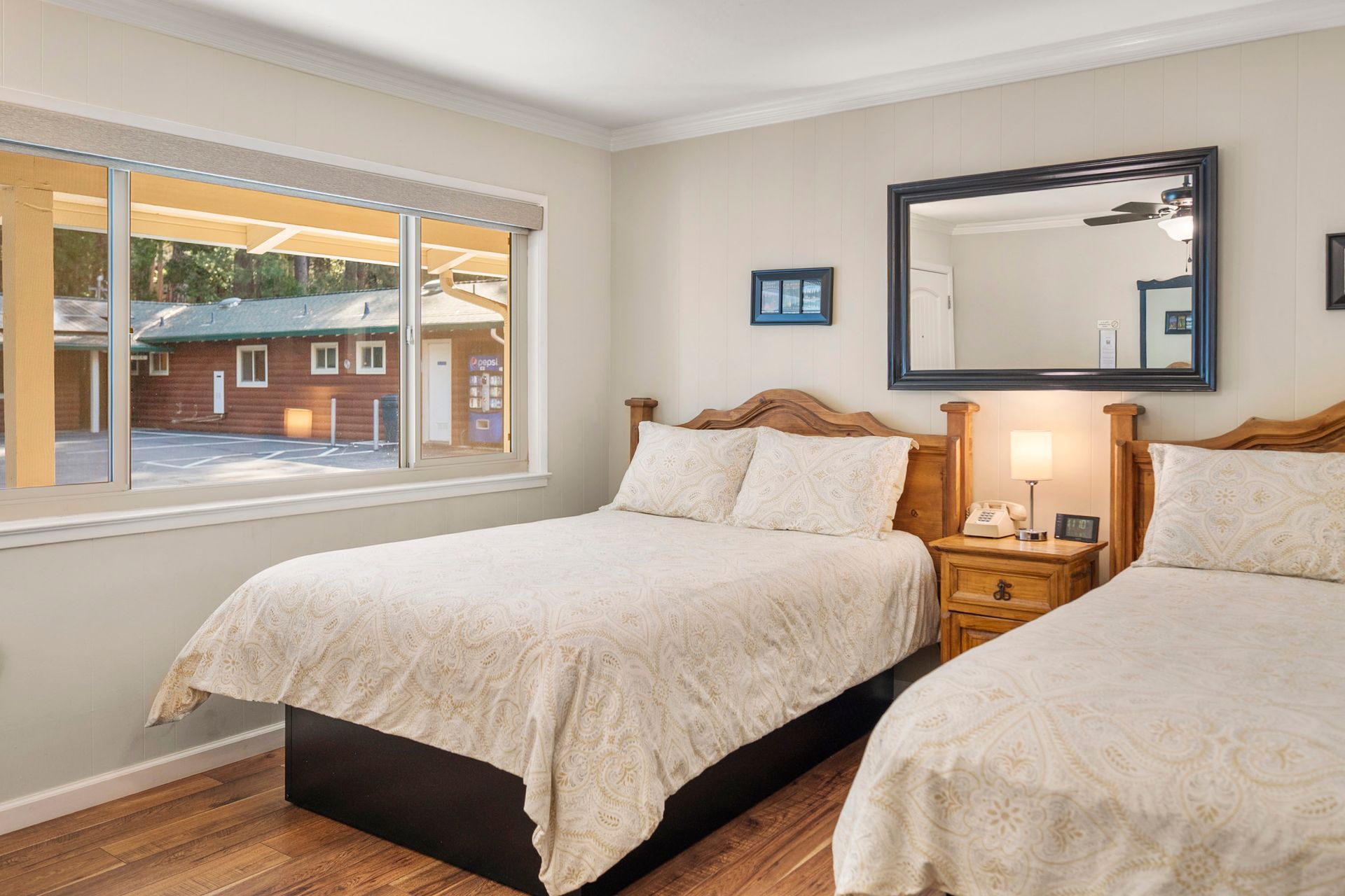 A hotel room with two beds with white patterned quilts, a nightstand with a lamp, and a large window looking outside.