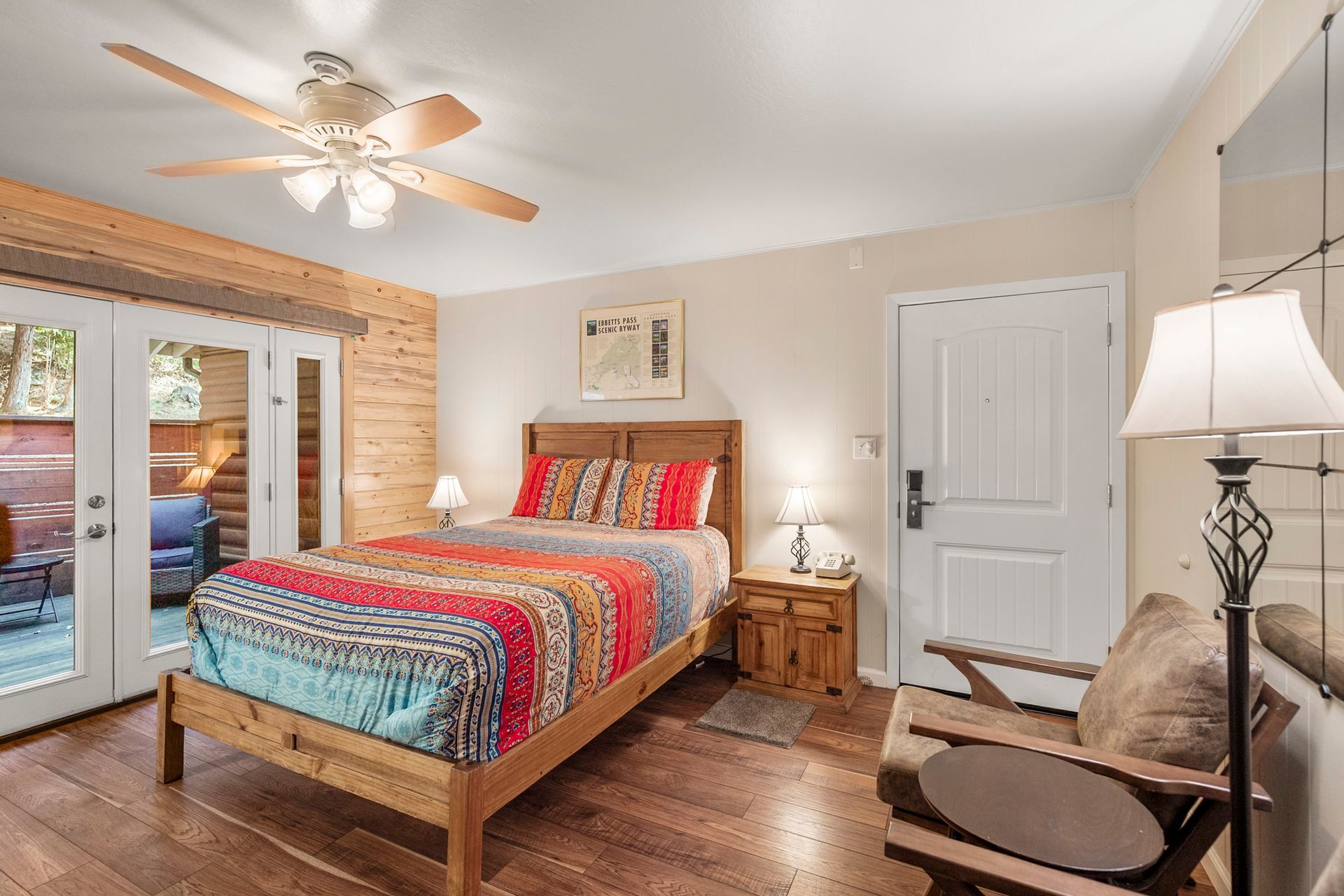 A furnished hotel room with a bed featuring a colorful patterned quilt, wooden headboard, ceiling fan, and arm chair.