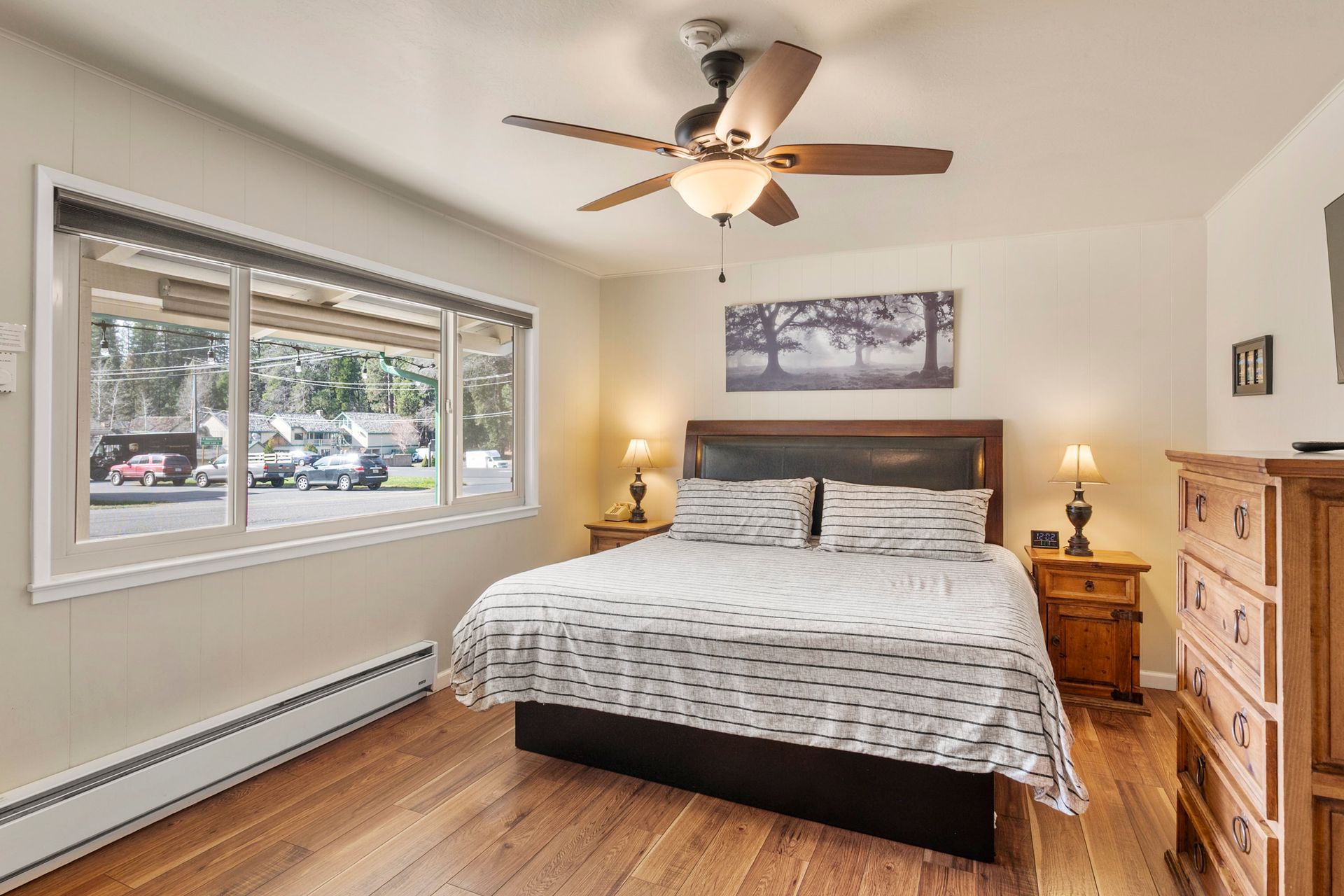 A bedroom with hardwood floors, a king bed with a patterned comforter, a ceiling fan, and a large window.