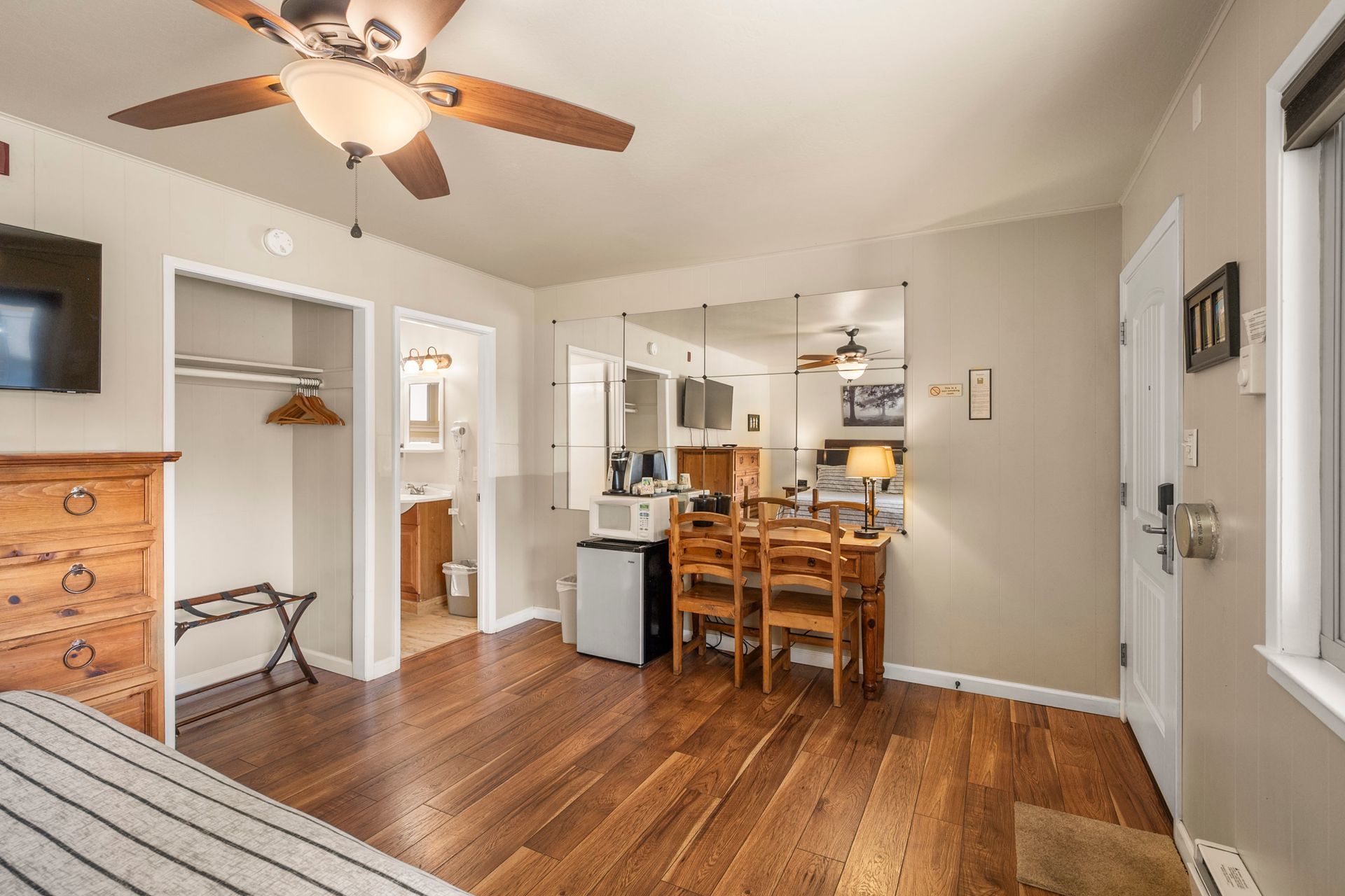 Hotel room interior featuring a wooden dresser, closet, small dining table with chairs, mini-fridge, and wood floors.