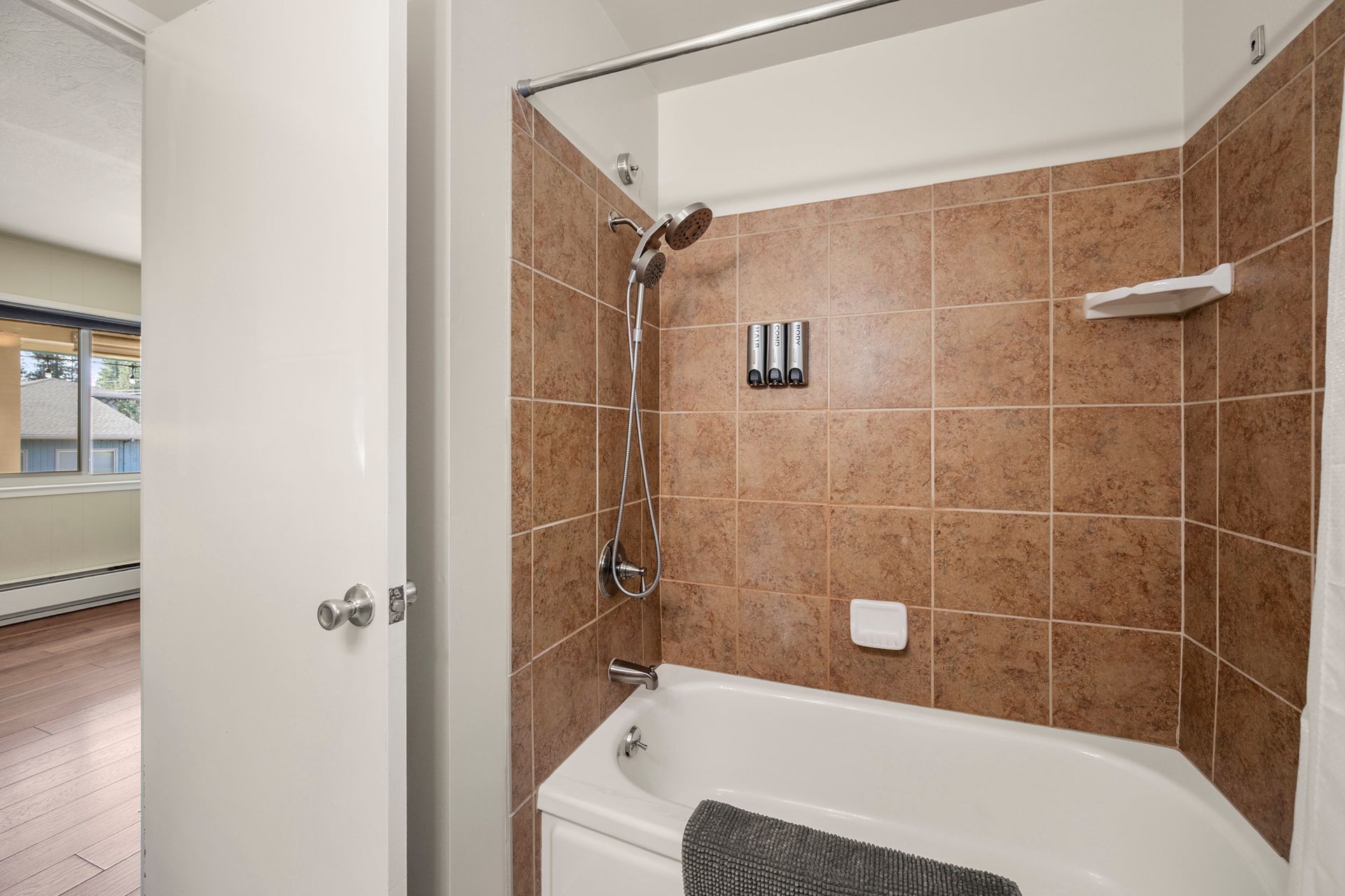 A bathroom with a white bathtub and shower fixture against brown tiled walls, next to an open door leading to a room.