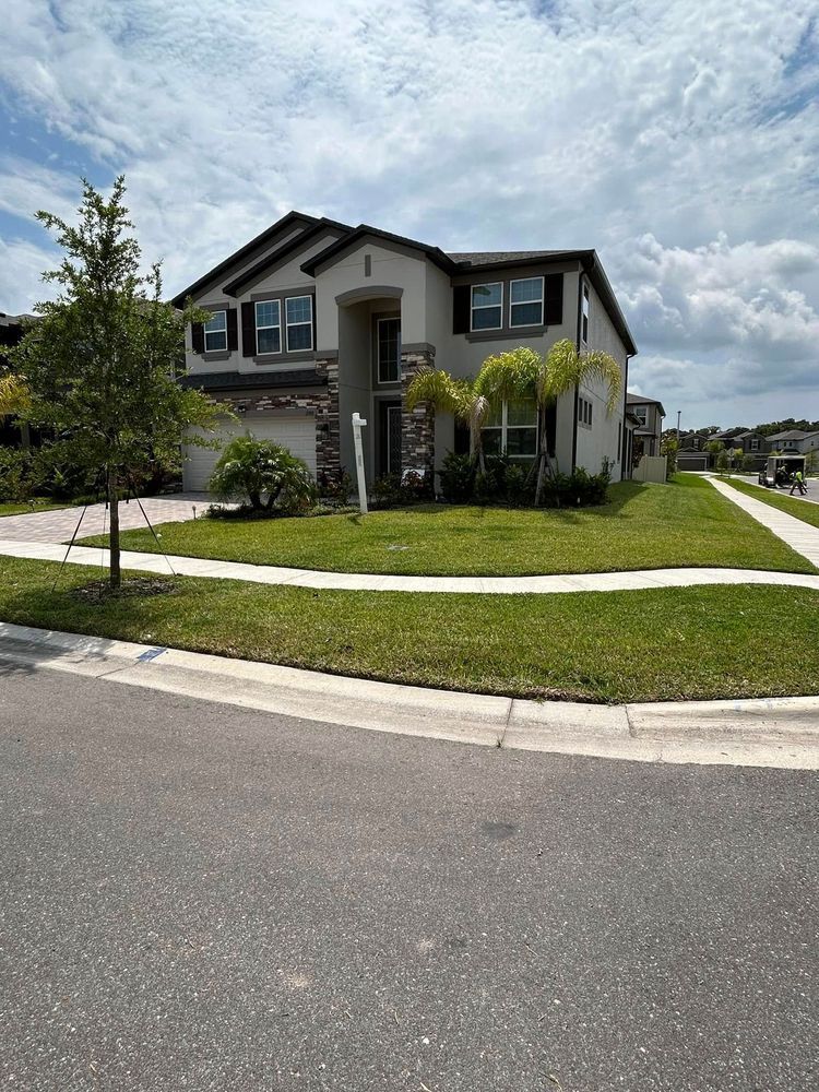 Two-story beige house with dark roof and trim, green lawn, in a suburban neighborhood under a cloudy sky.