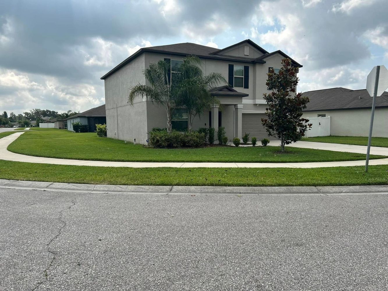 Two-story house with beige siding, dark roof, and manicured lawn on a suburban street.