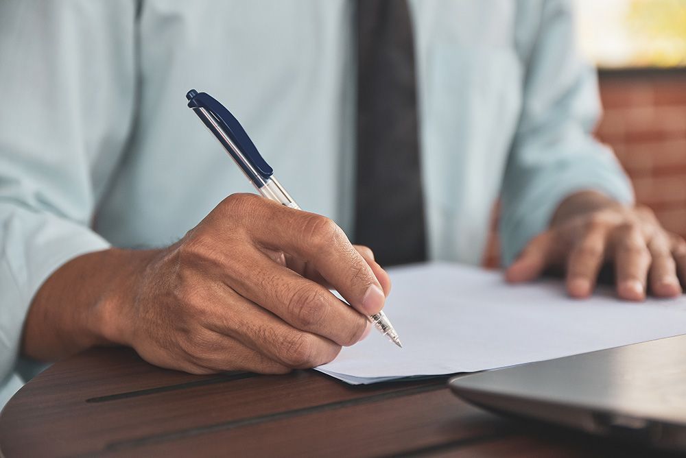 Person in a light blue shirt and black tie writing on a white paper with a pen, wooden table.
