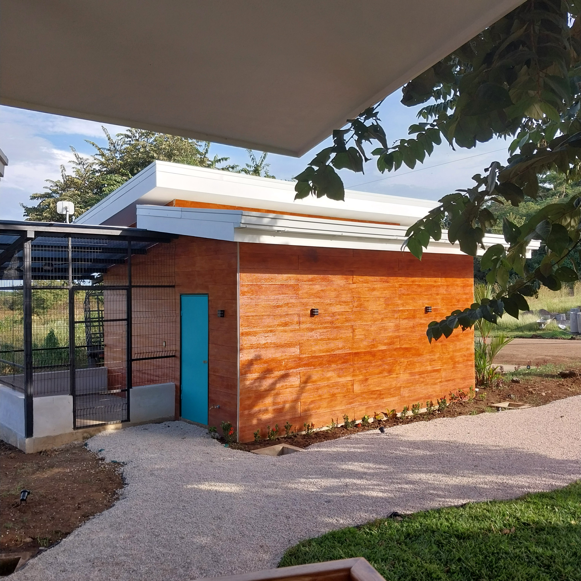 Brick building with blue door and gravel path. A pergola and foliage are in view.