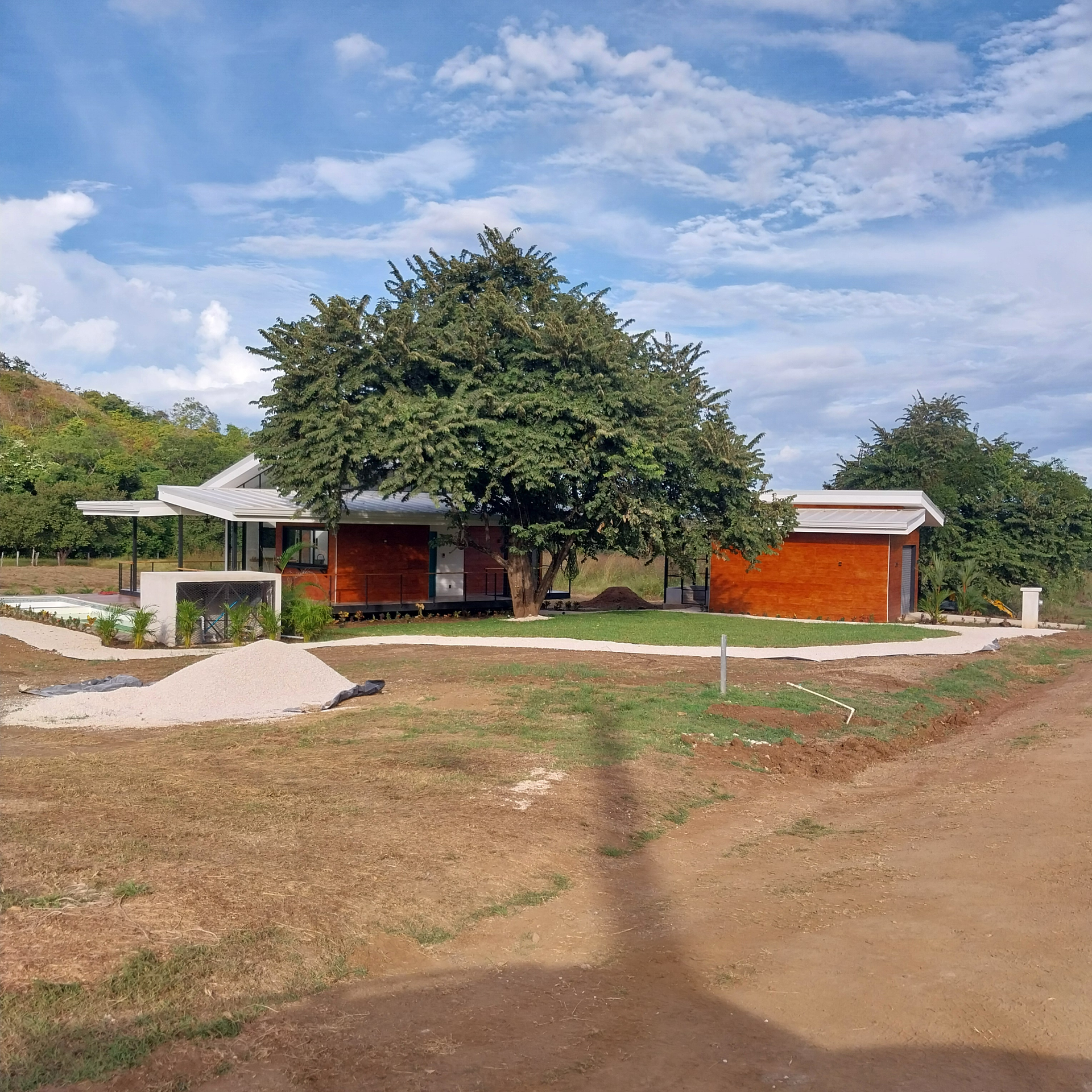 Modern, single-story house with wood siding, under a blue sky, surrounded by grass and a dirt road.