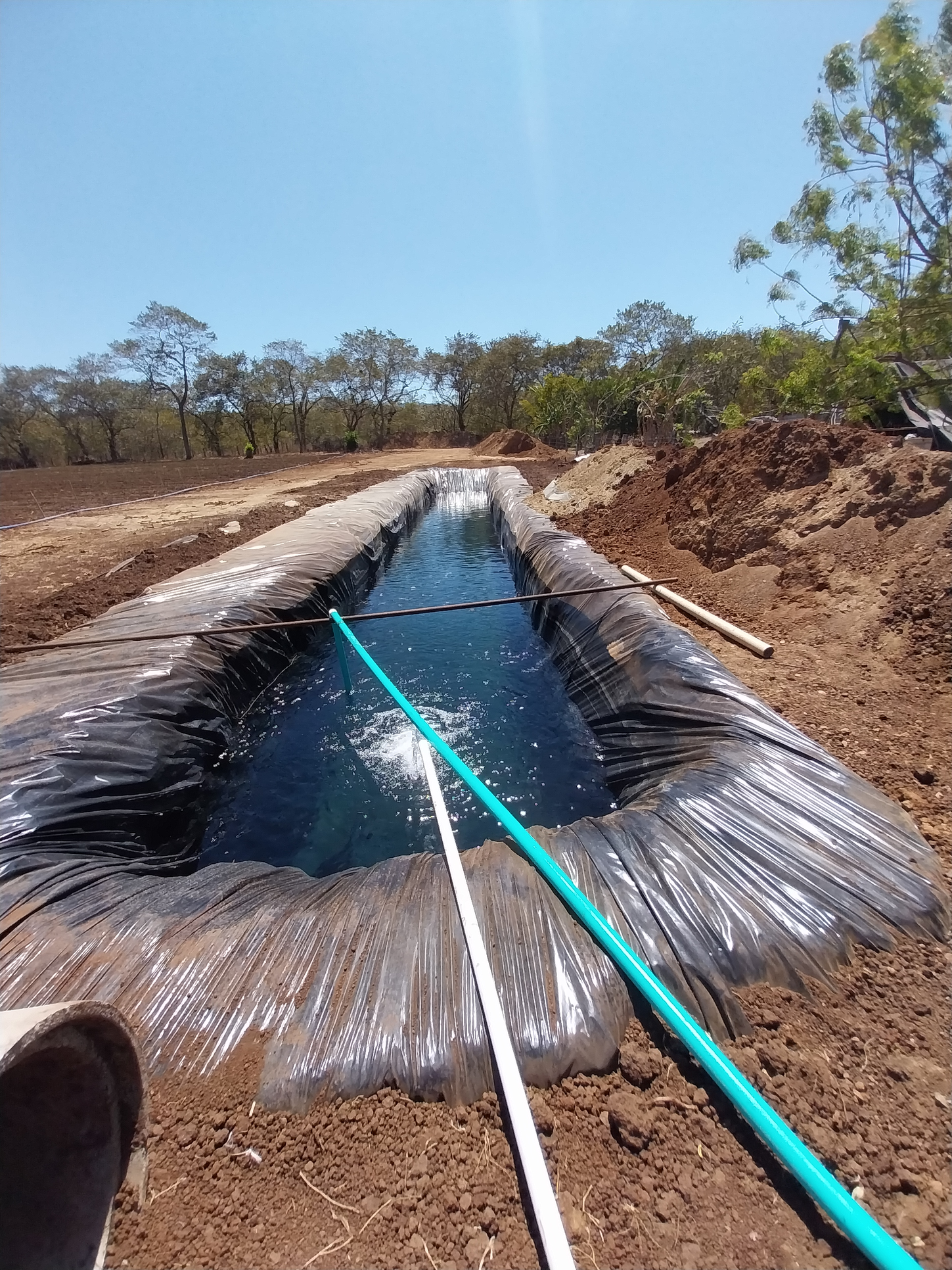 Water-filled, black-lined irrigation trench in a dry, outdoor setting under a clear blue sky.