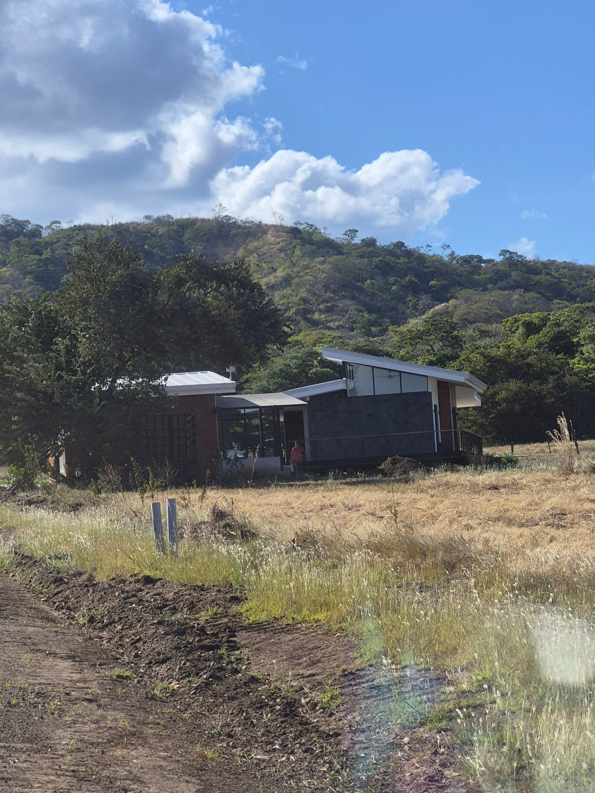 House in a field with a mountain backdrop, under a cloudy blue sky.