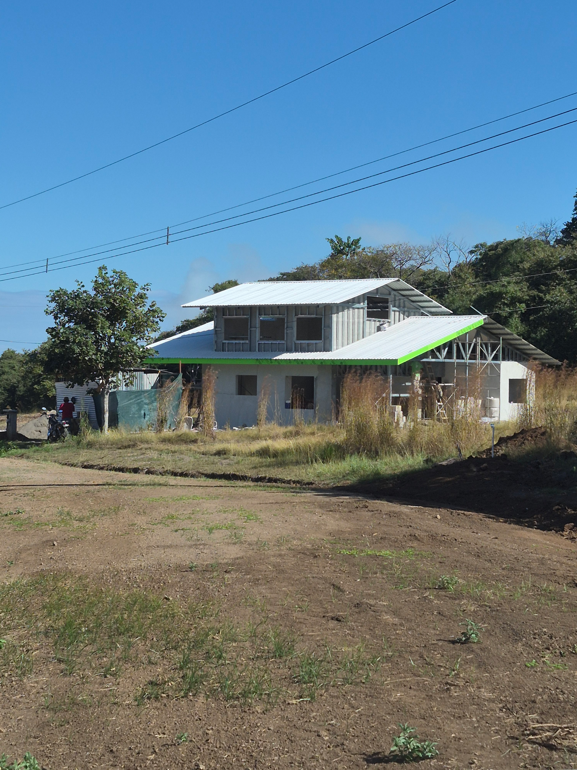 Two-story house with gray walls, green trim, and a metal roof against a blue sky, trees in the background.