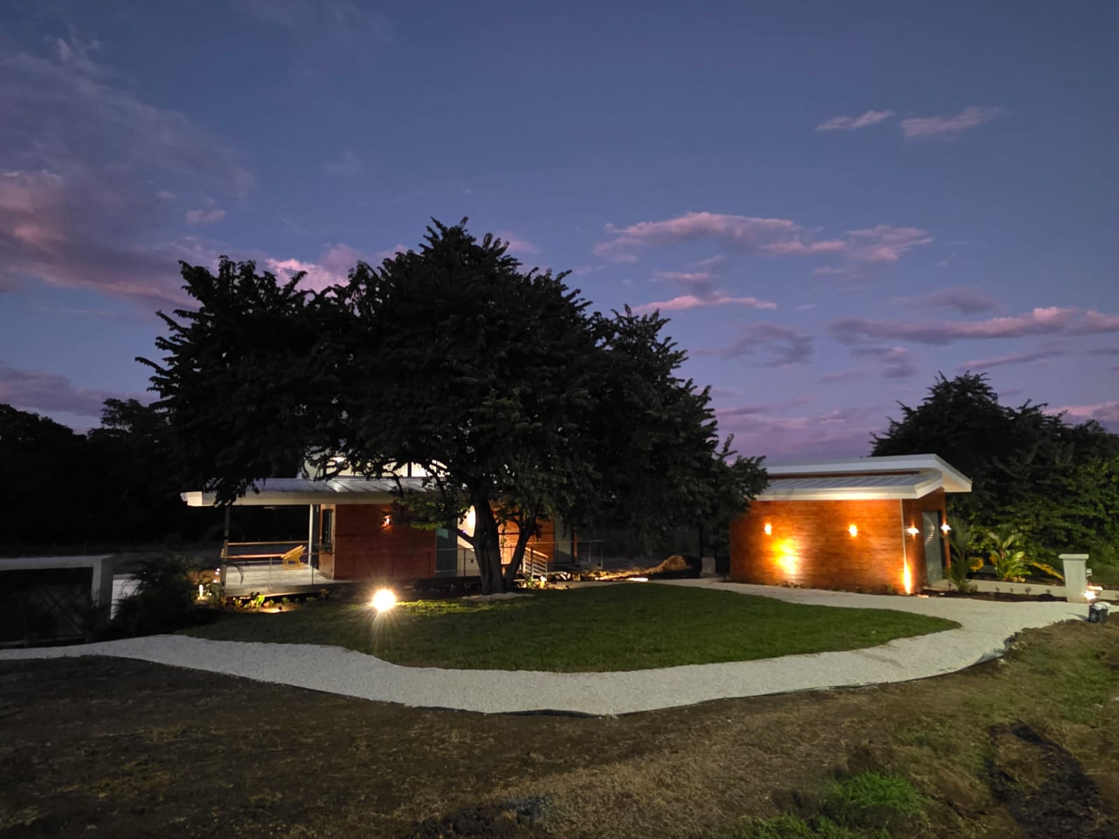A modern, low house with a lighted pathway and mature tree at dusk; sky with pink and purple hues.