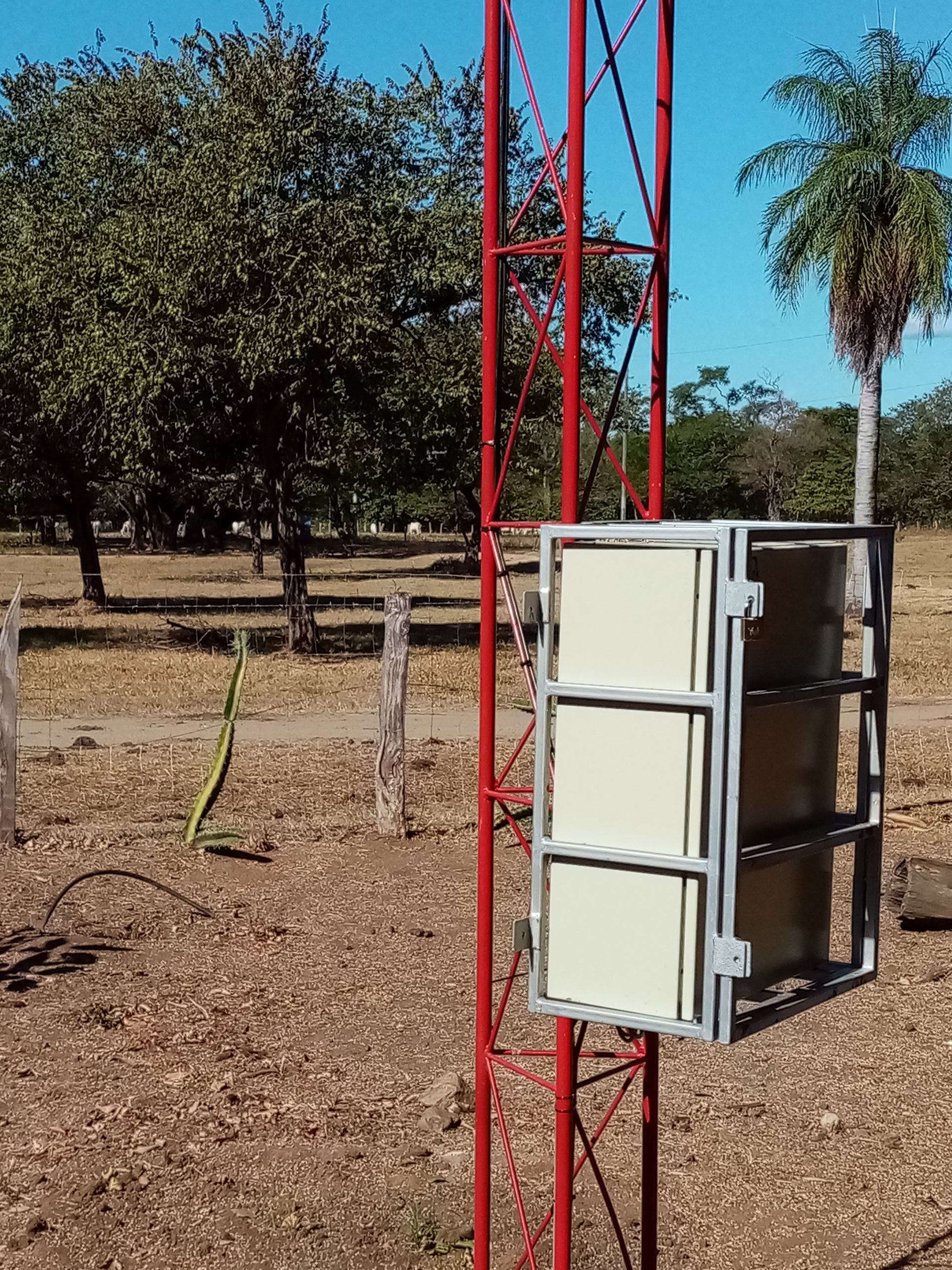 Red lattice tower with a white, rectangular enclosure mounted to it, outdoors.