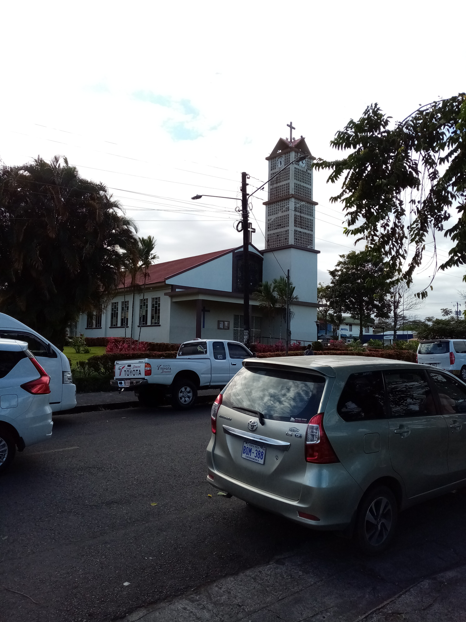 Church with a white bell tower and cross, surrounded by parked cars and trees under a cloudy sky.