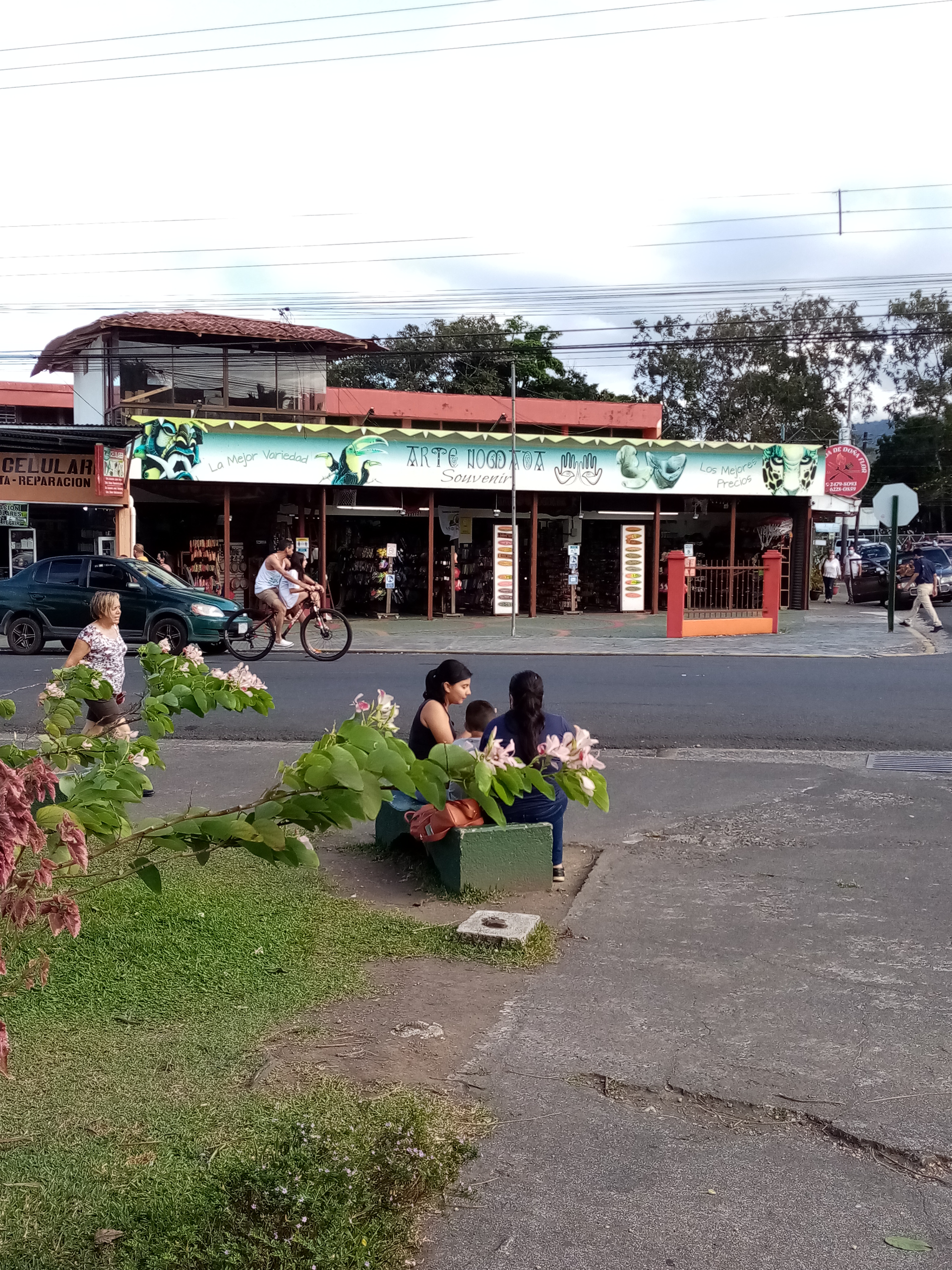 People sitting on a bench with flowers, street view of a store with a cyclist and other pedestrians.