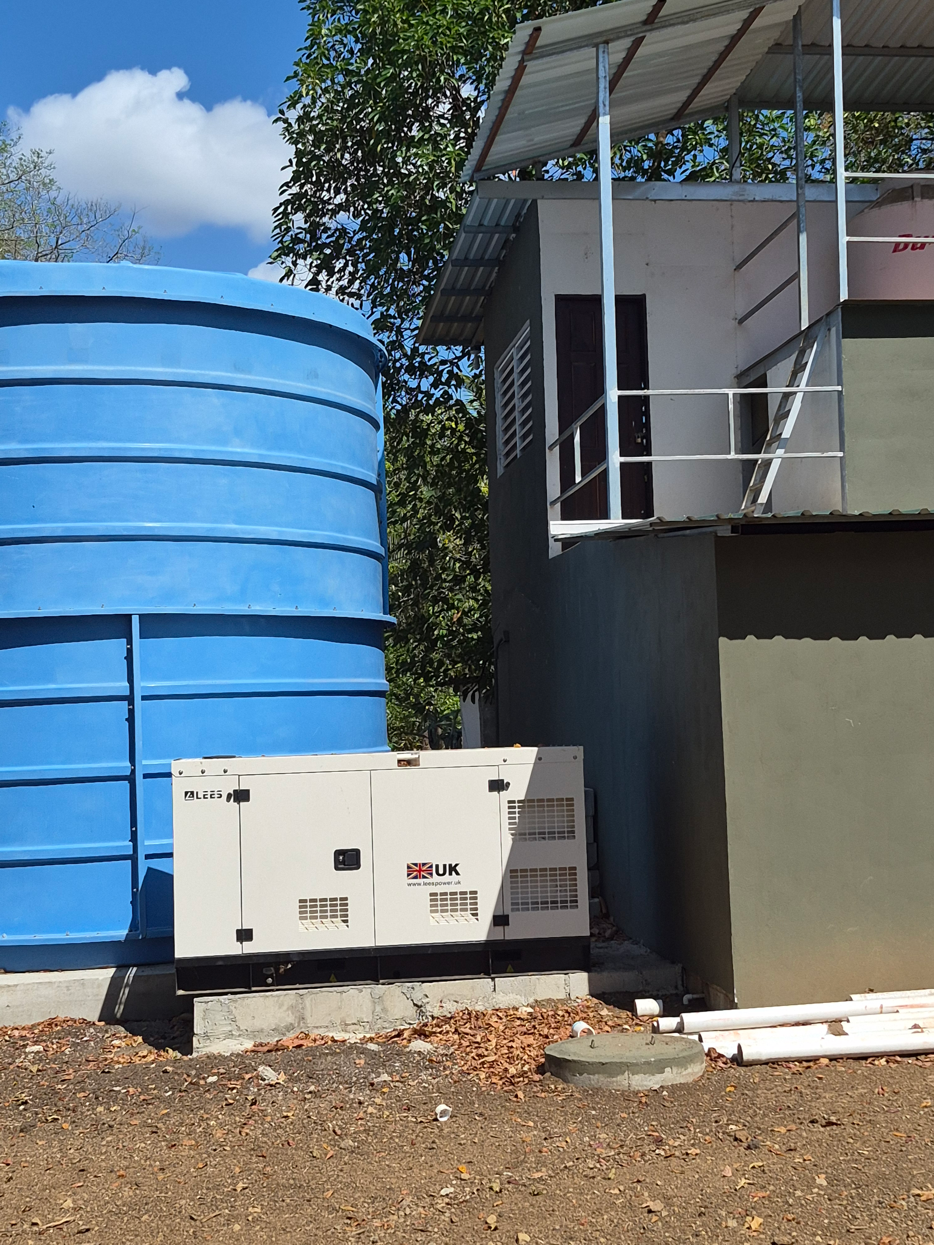 Blue water tank, white generator, and small building in outdoor setting with blue sky.