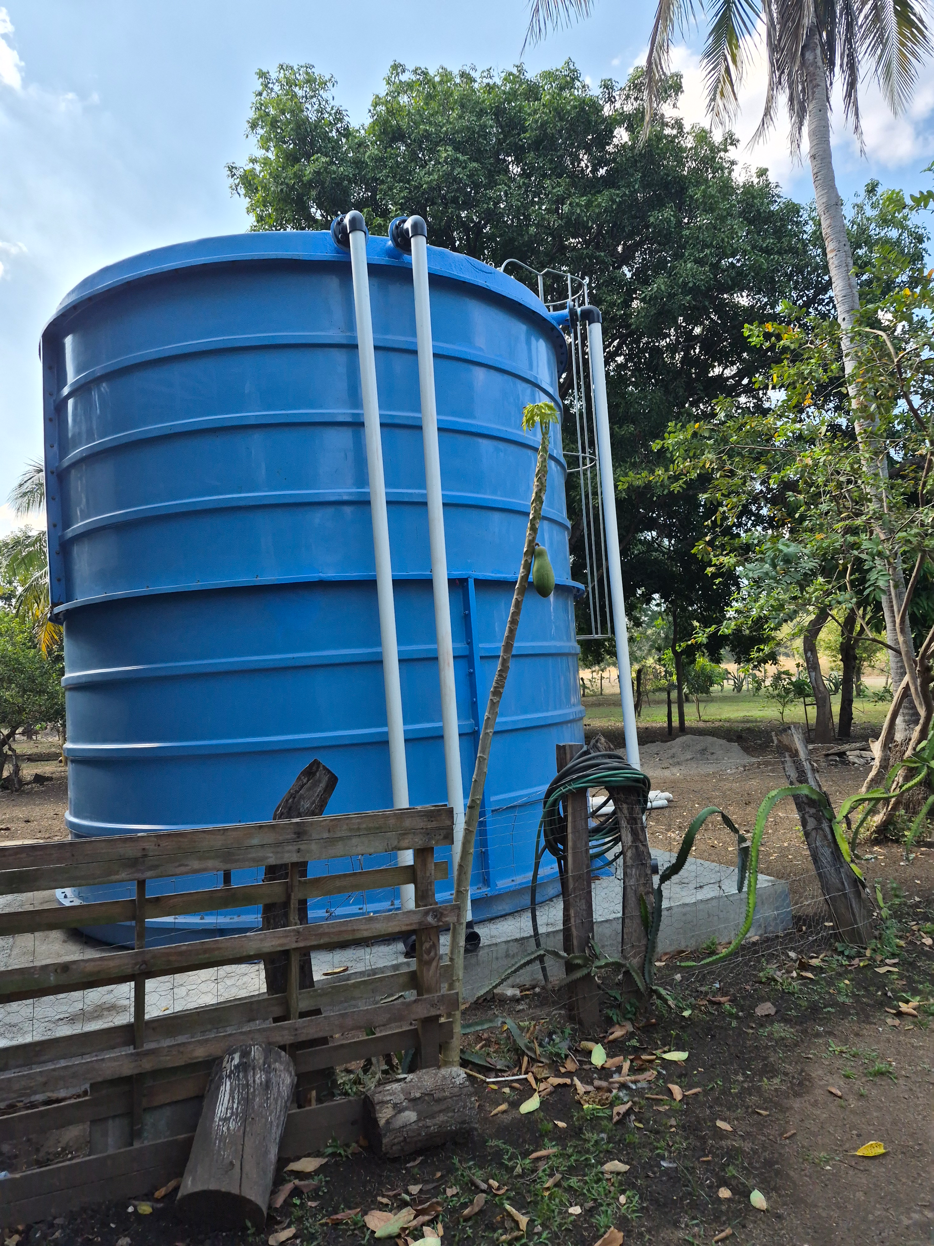 Blue water tank with white piping and ladder, outdoors near trees and a wooden fence.