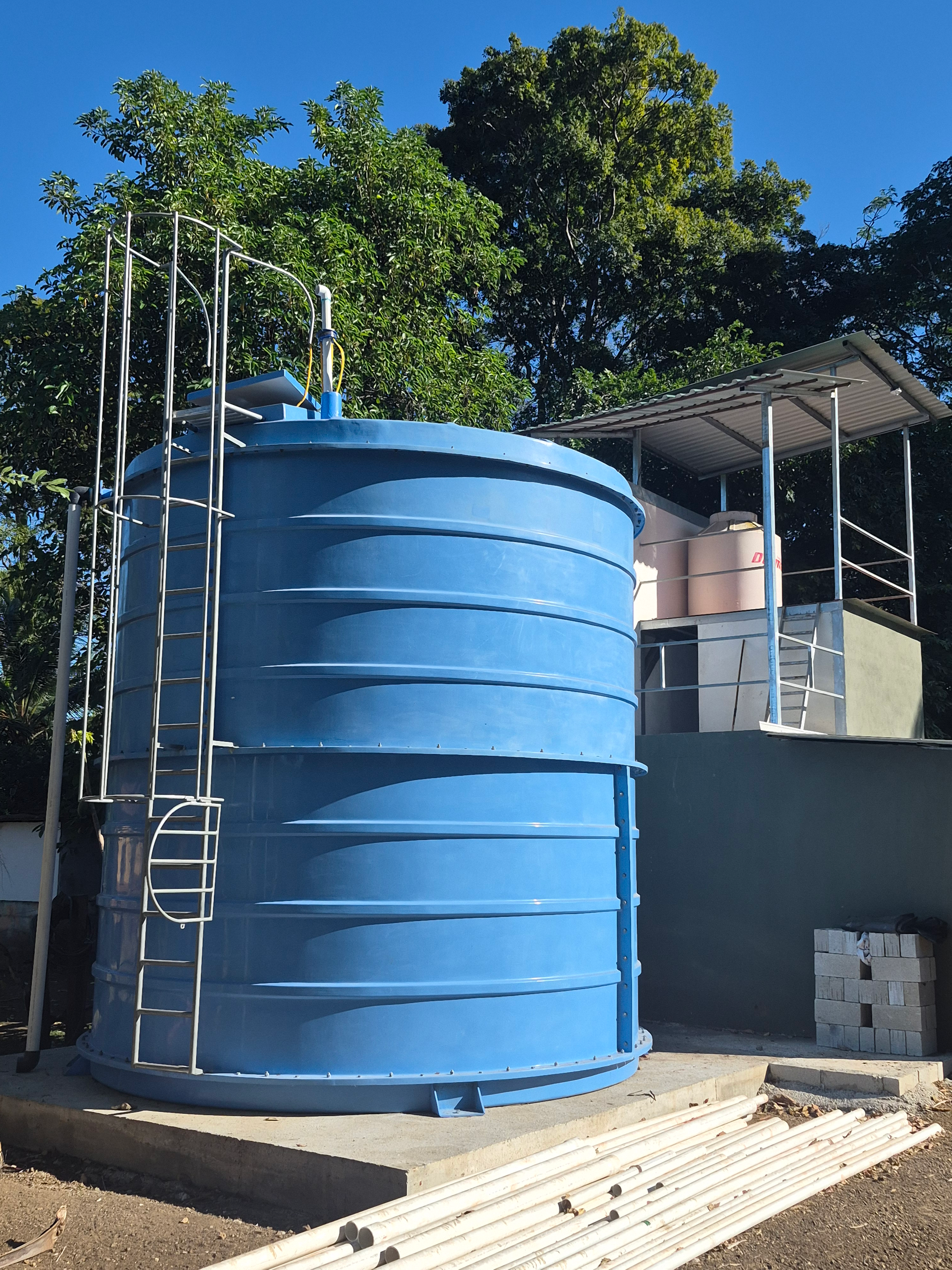 Blue water tank with ladder, near a shelter, set against trees and a blue sky.