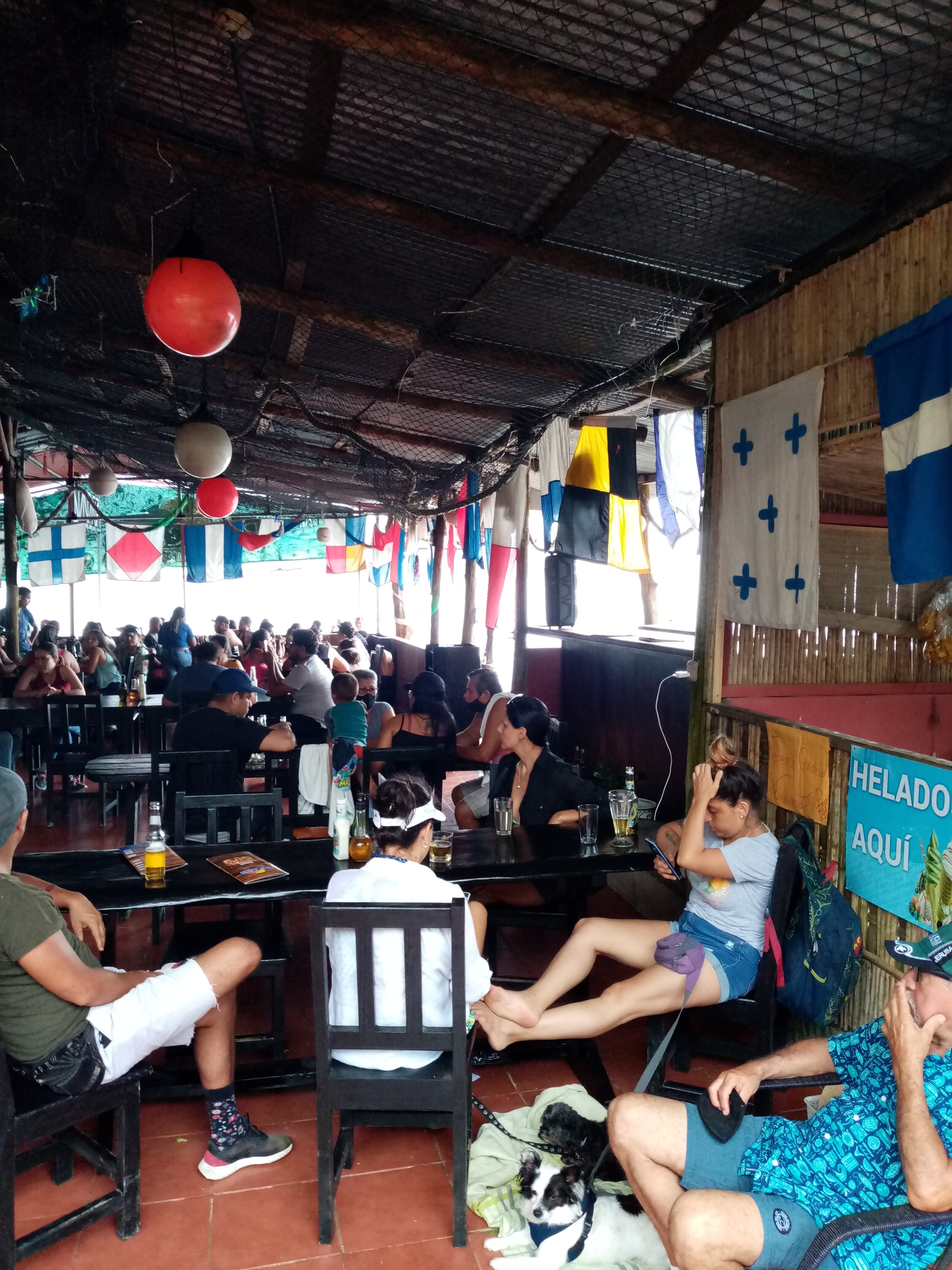 People in a casual outdoor cafe, tables, chairs, flags, red and white lights, blue and white flag.