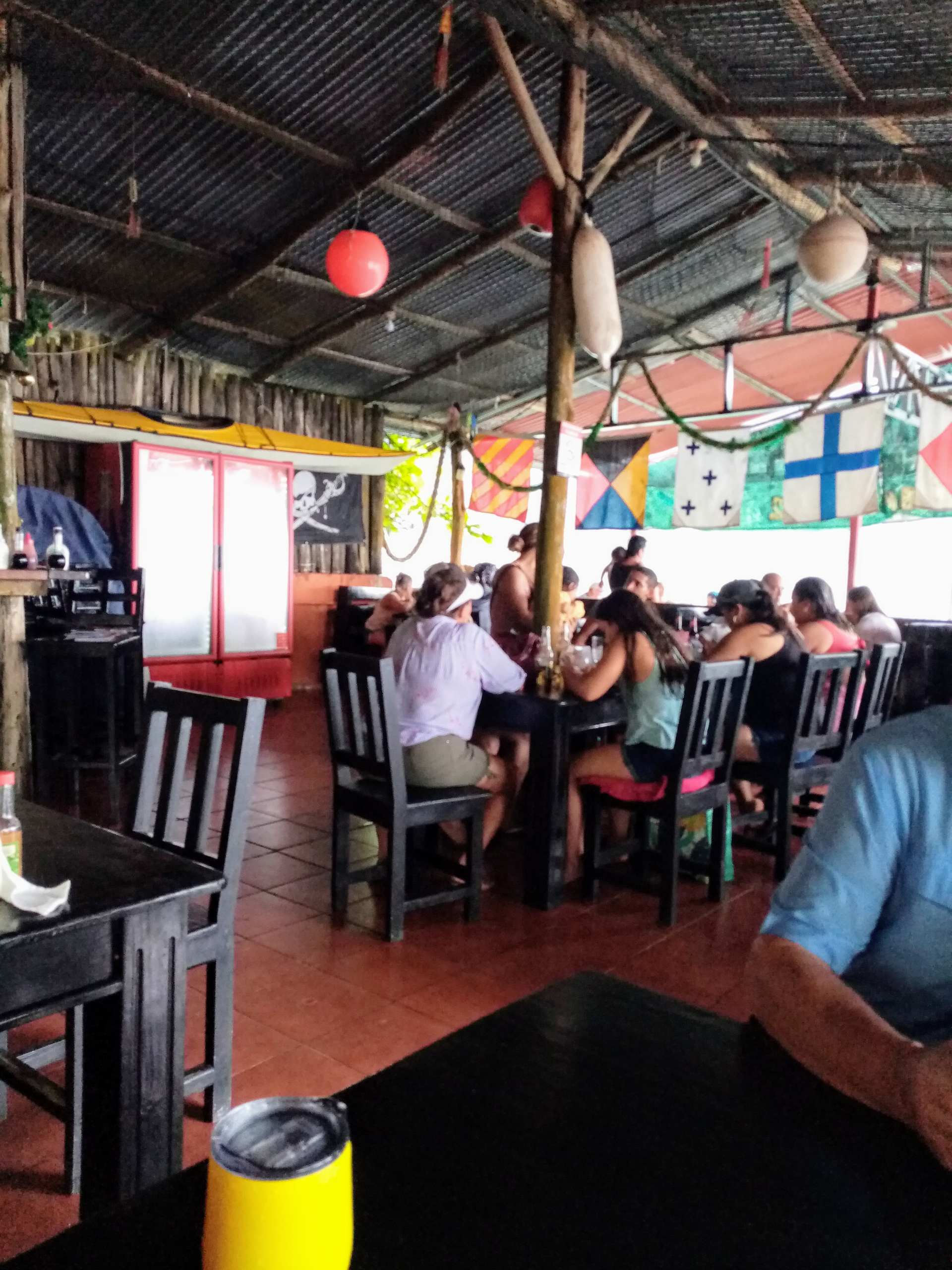 Restaurant interior with seated patrons. Tables and chairs. Nautical decor, corrugated metal roof, red floor.