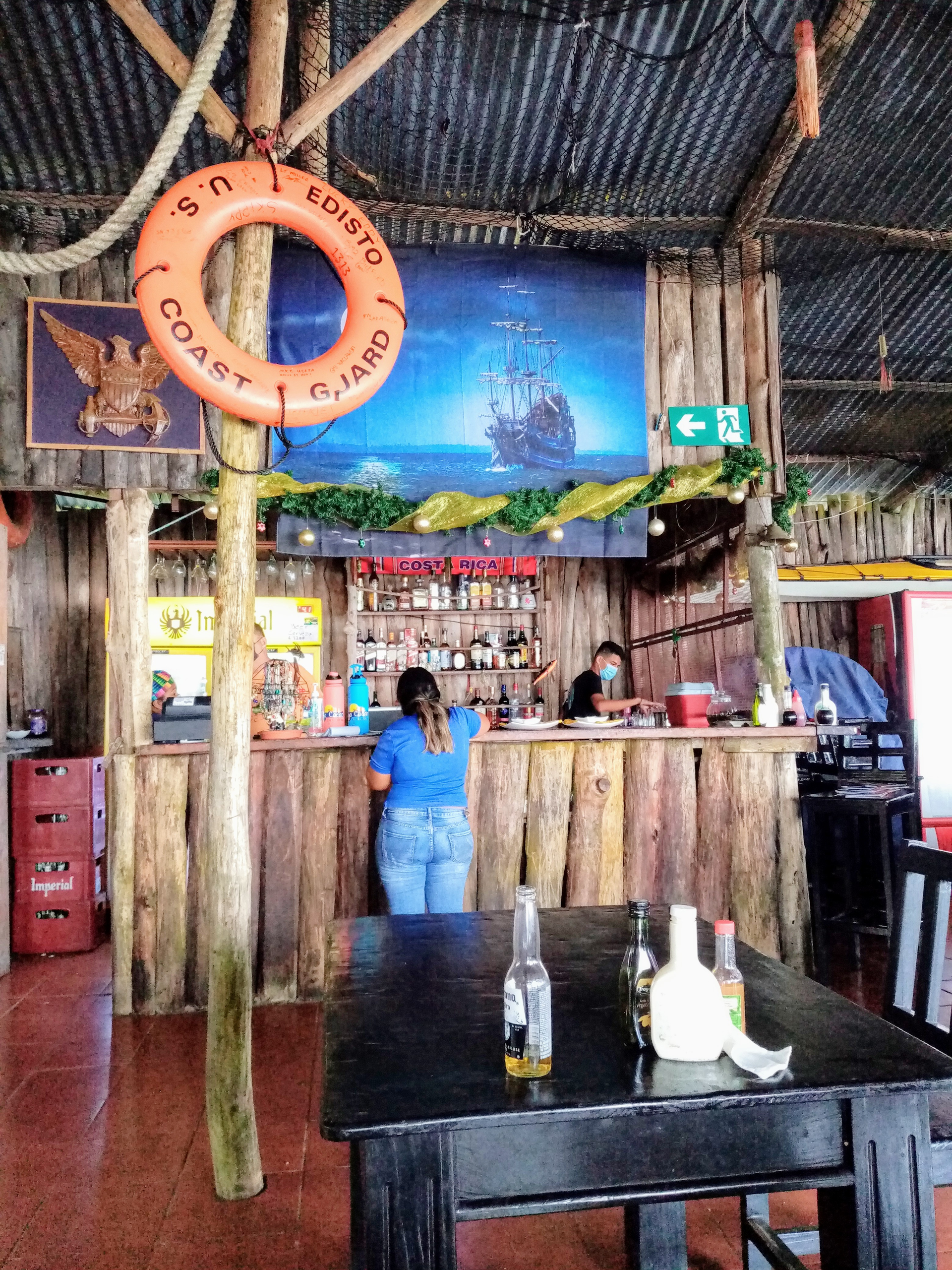 A rustic bar with two people behind it, a ship painting, and a Coast Guard buoy.