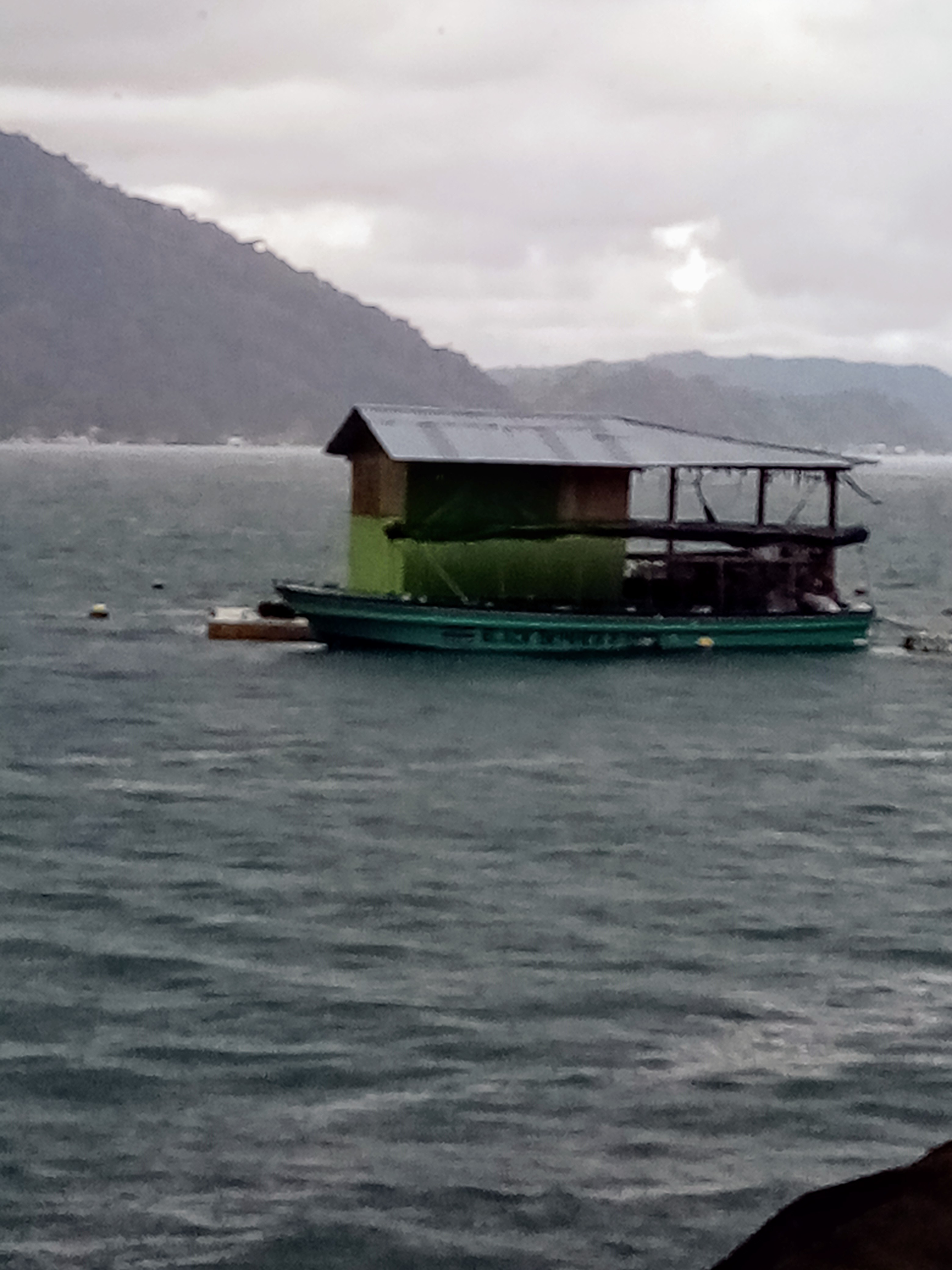 Green floating structure with a building on top, in choppy water near a mountain.
