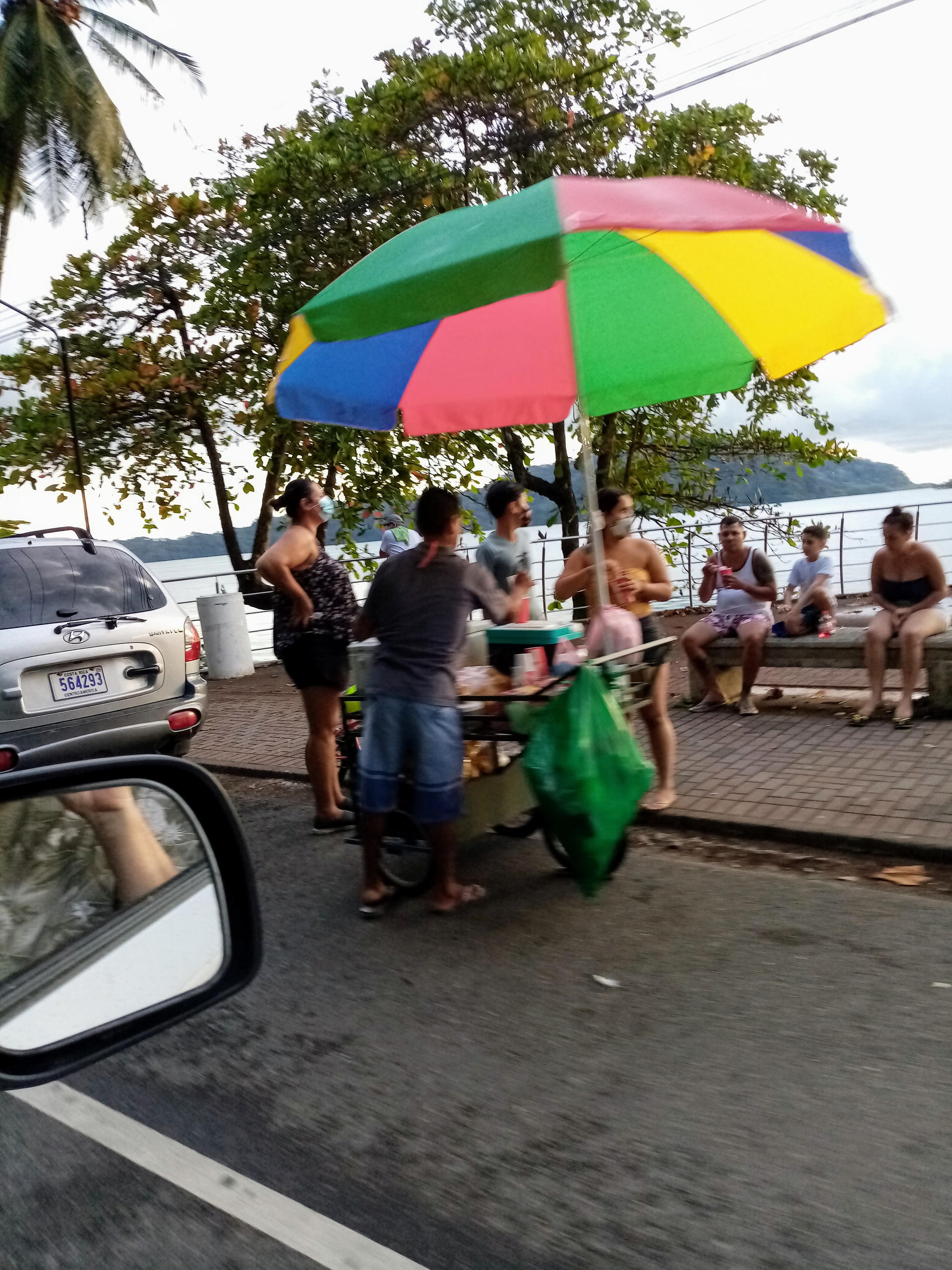 Street vendor with colorful umbrella, selling food by the water. People are gathered around the cart.