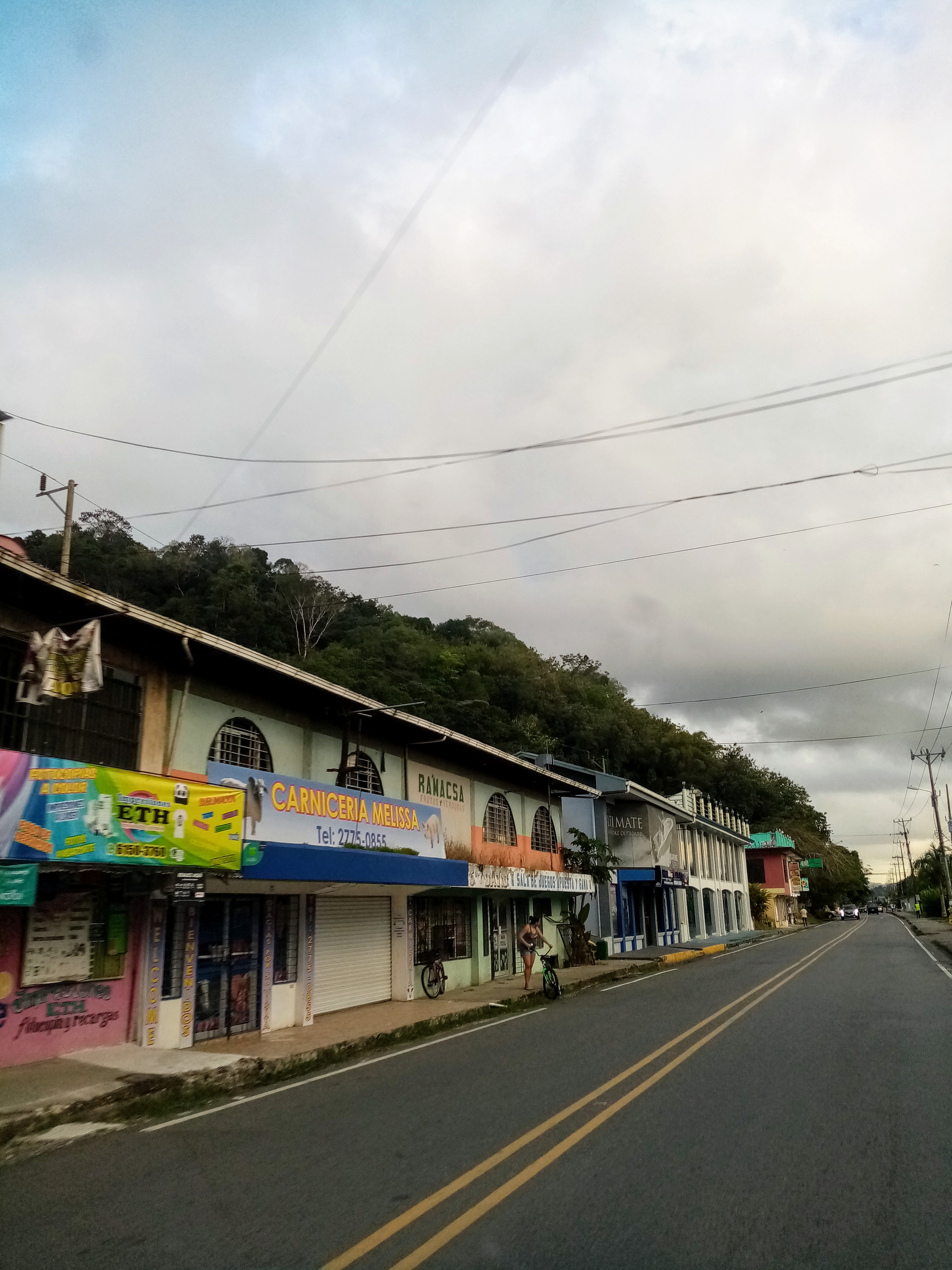 Shops line a road, with a hillside in the background under a cloudy sky.