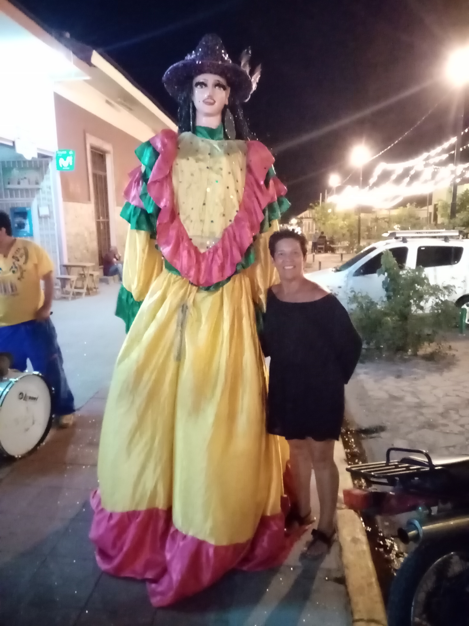 Woman stands next to a tall costumed figure in a yellow dress. Night setting, street, drums, and car.