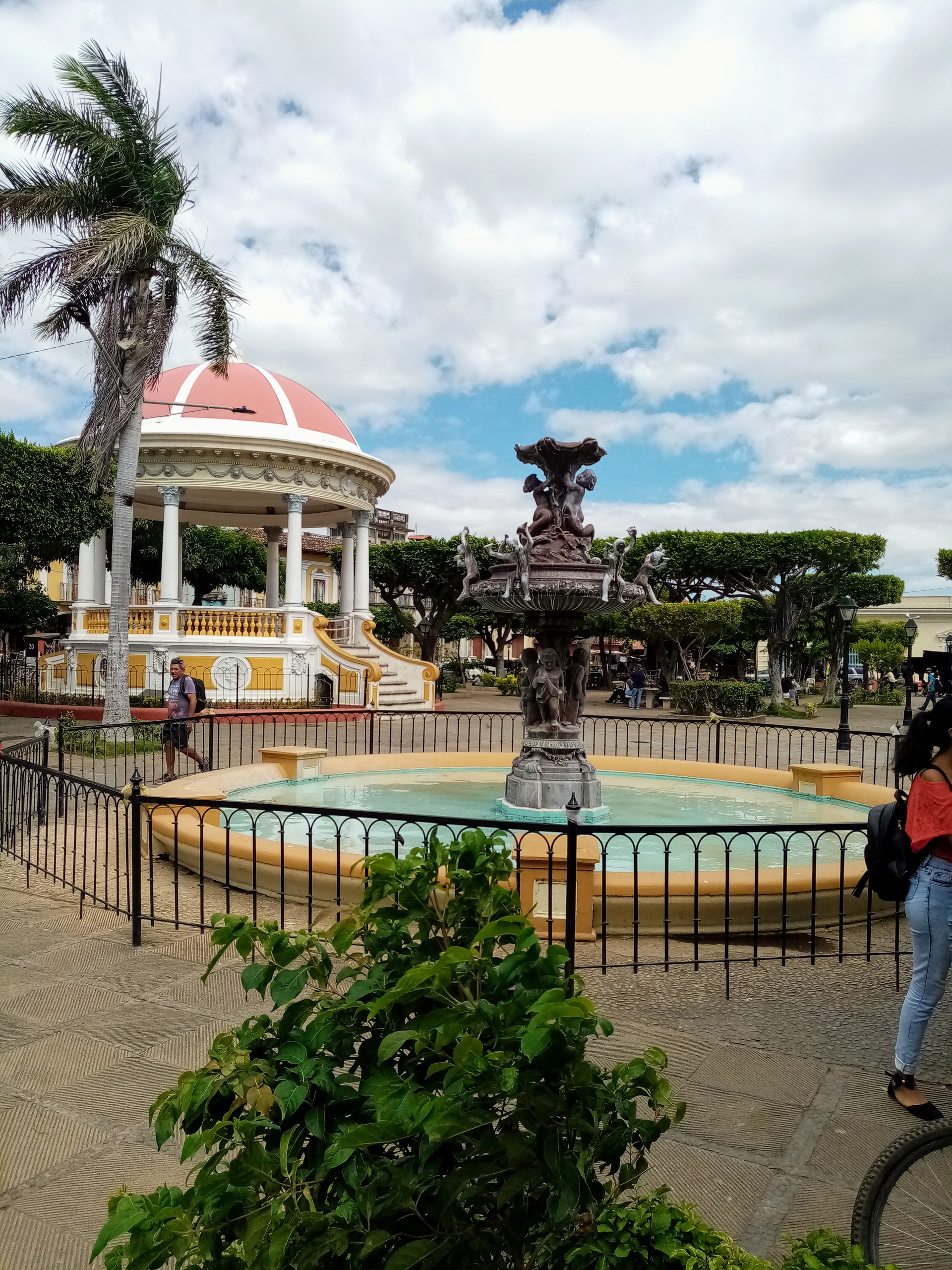 Fountain and gazebo in a town square, blue sky with clouds.  Person walks past gazebo.