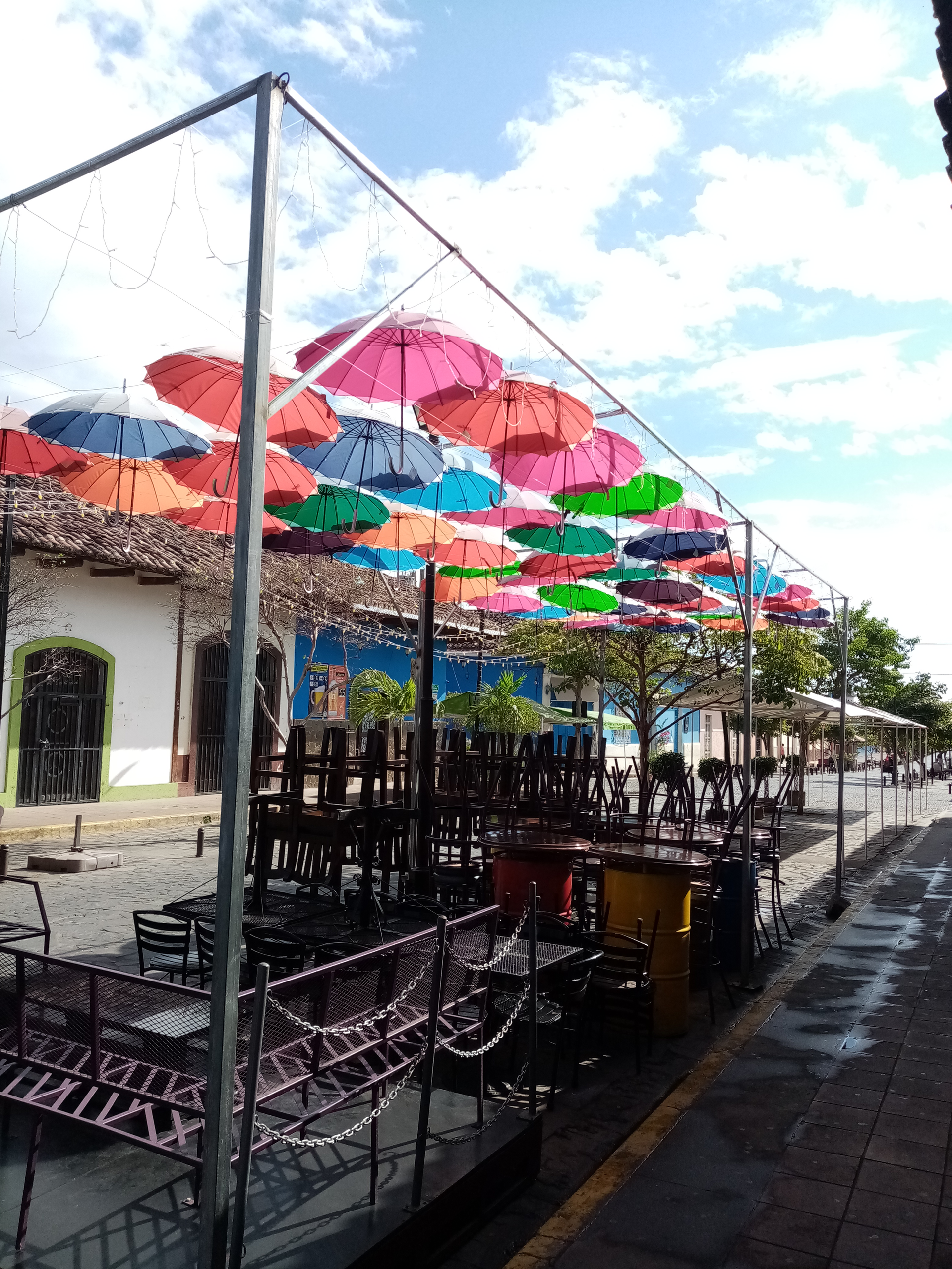 Row of colorful umbrellas strung over outdoor seating area in a town. Bright sky.