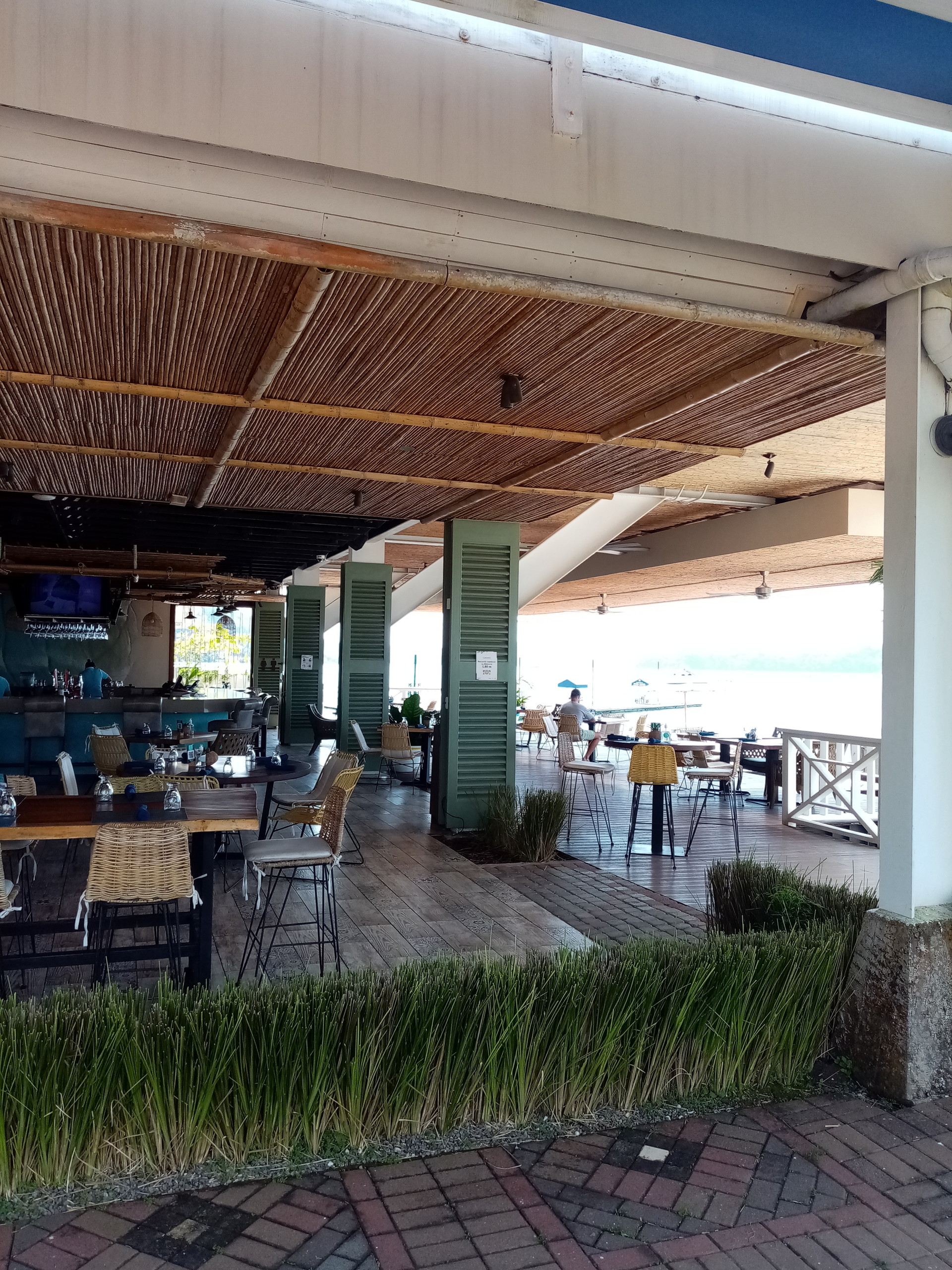 Outdoor restaurant with wooden deck, tables, and a bar under a woven bamboo ceiling; water in the distance.