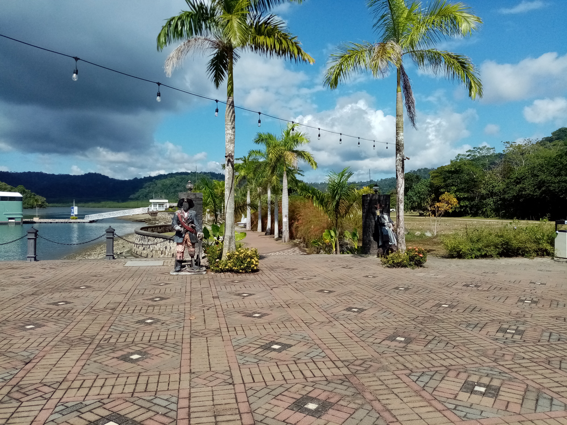 Brick walkway with palm trees, waterfront, cloudy sky. Two people walking, string lights overhead.