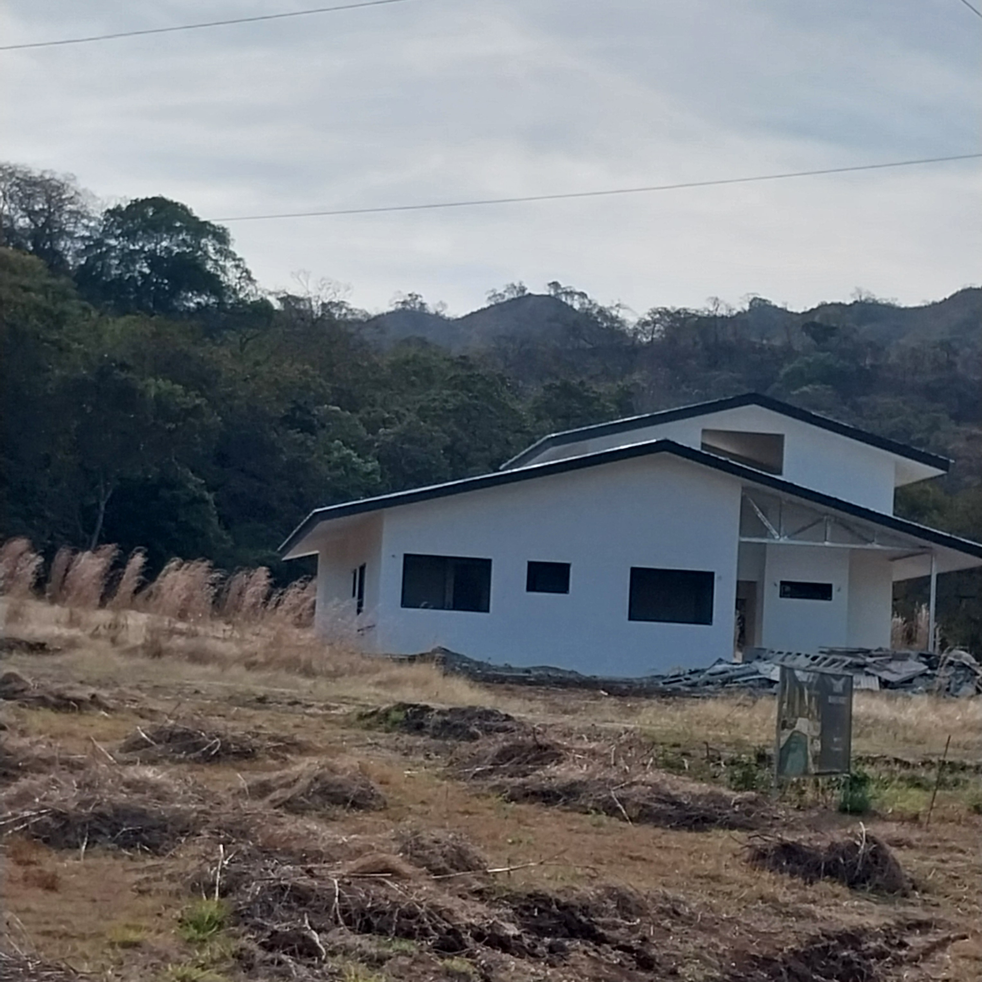 White house under construction in a dry, grassy field with a backdrop of forested hills and a cloudy sky.