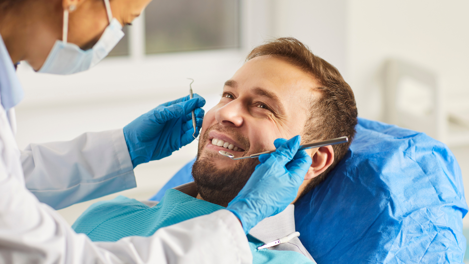 Dentist examining a patient's teeth with dental instruments; blue gloves, mask, and dental chair cover.