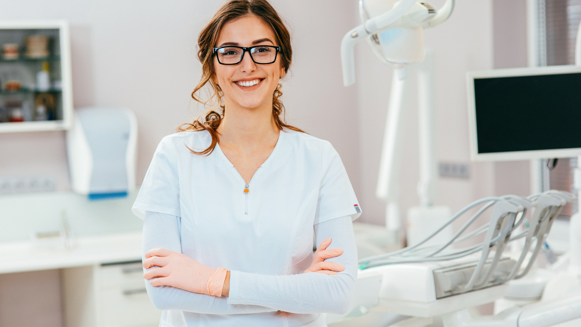 Woman in white scrubs and glasses smiles in a dental office.