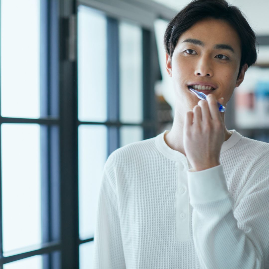 Man brushing teeth near a window, wearing a white long-sleeved shirt.