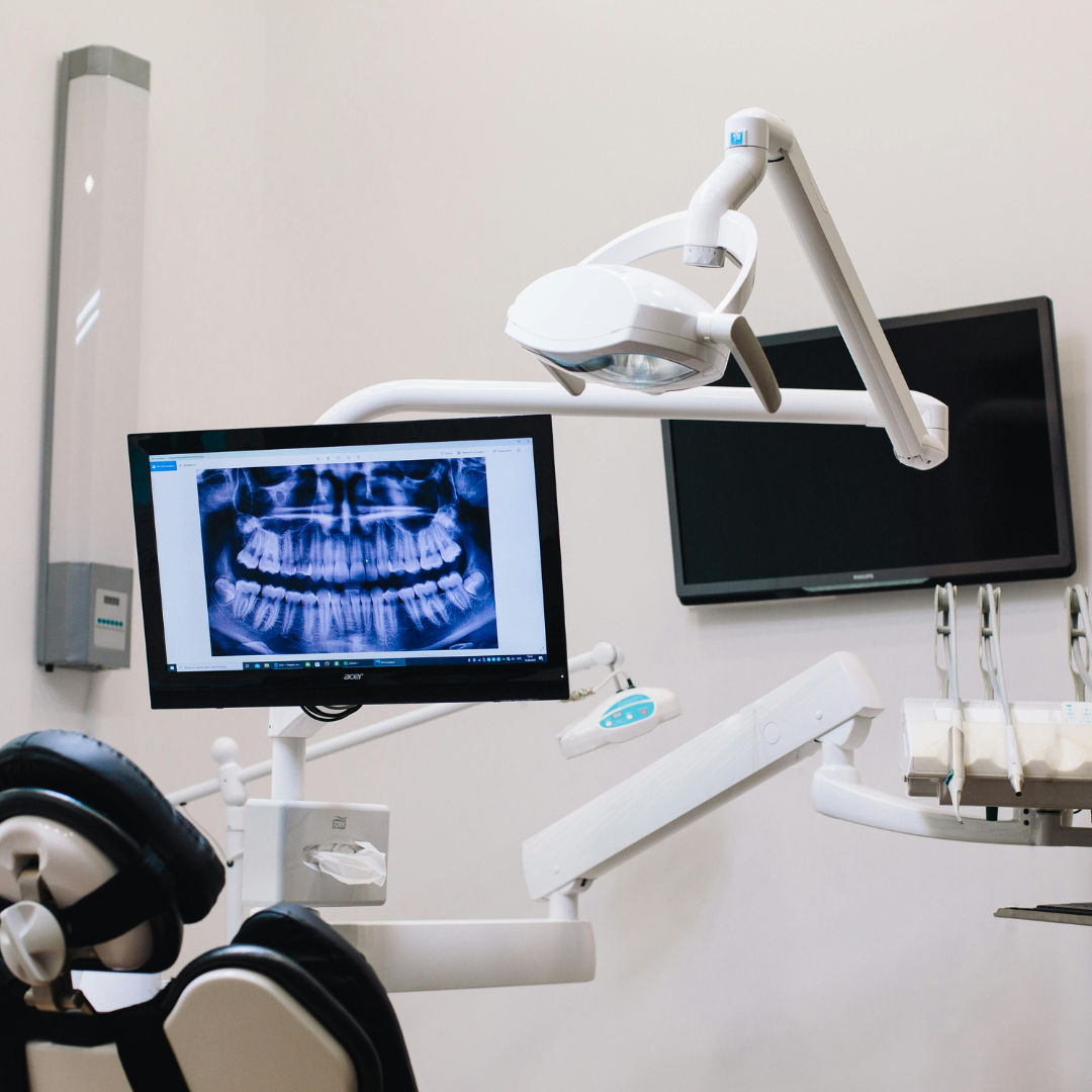 Dental office interior with patient chair, monitor showing dental X-ray, examination light, and equipment.