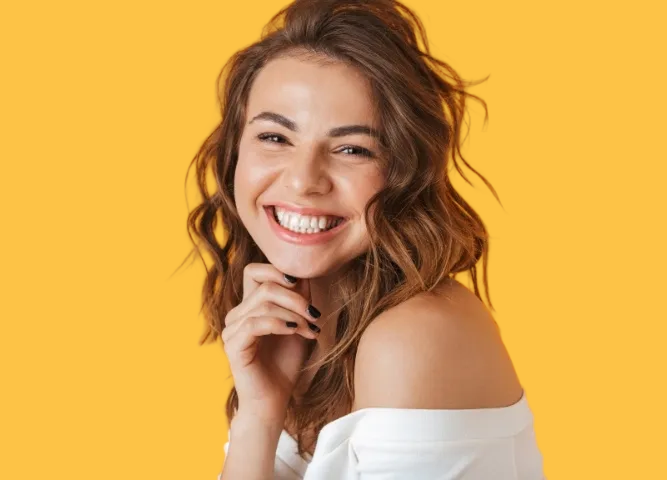 Woman smiling, hand near face, wearing white top, against yellow background.